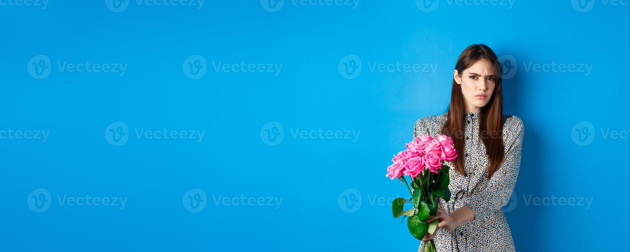 Valentines day concept. Displeased young woman rejecting flowers gift, frowning and grimacing, refusing take roses, standing on blue background photo