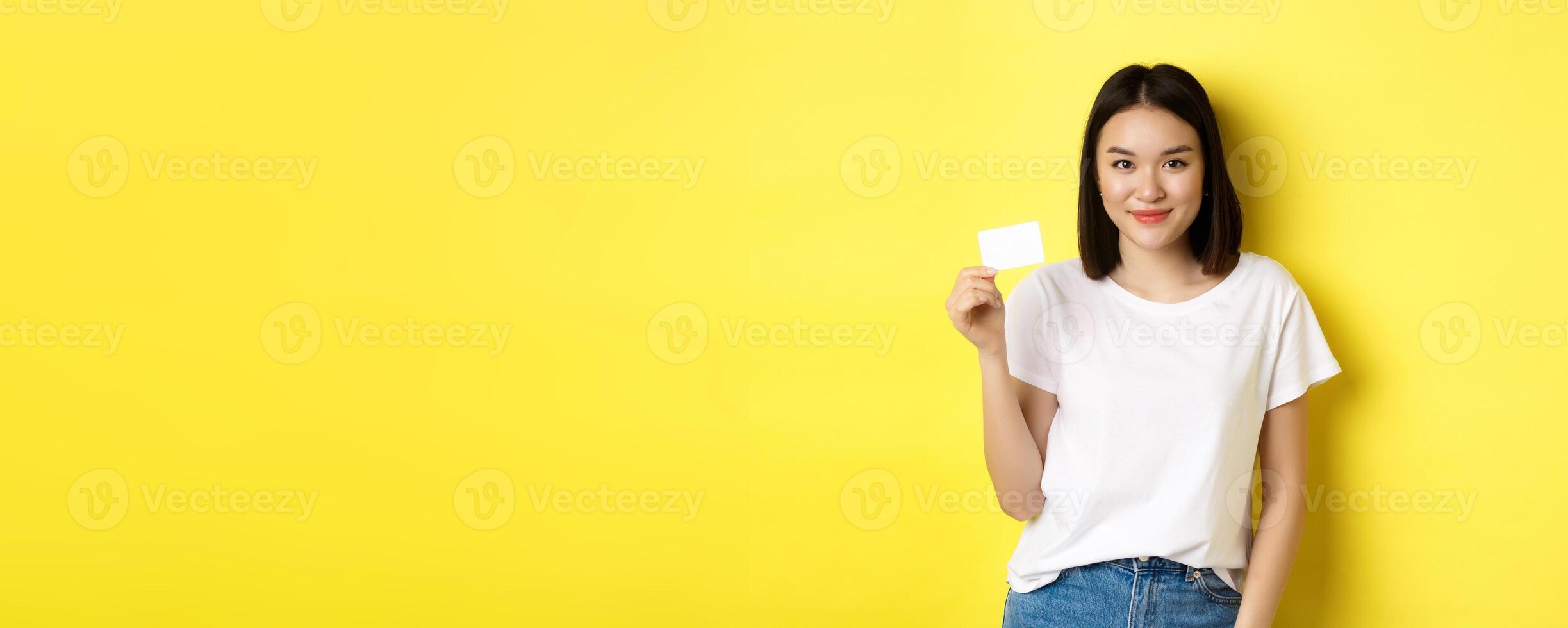 Young asian woman in casual white t-shirt showing plastic credit card and smiling at camera, yellow background photo