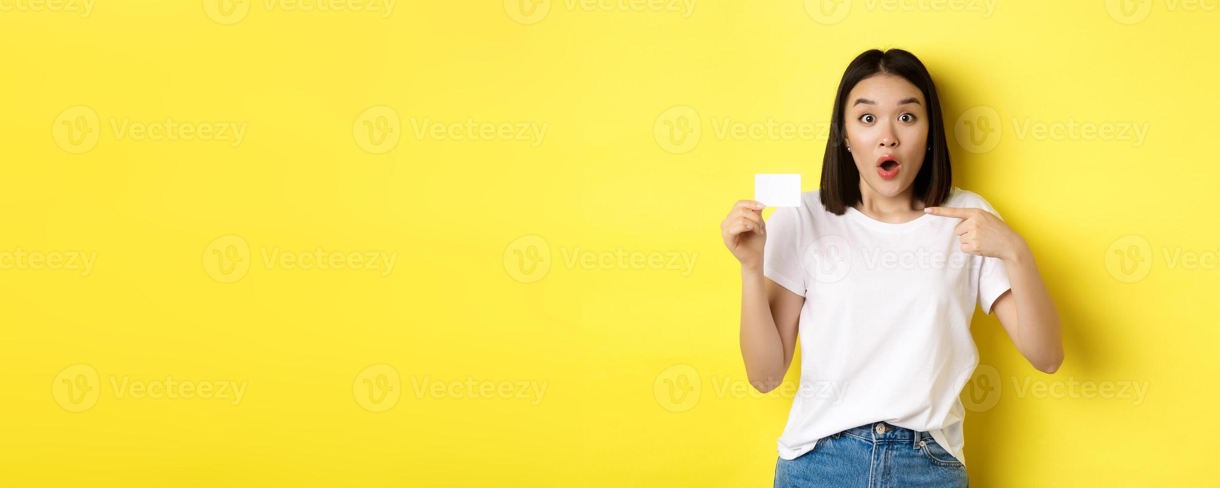 Young asian woman in casual white t-shirt showing plastic credit card and smiling at camera, yellow background photo