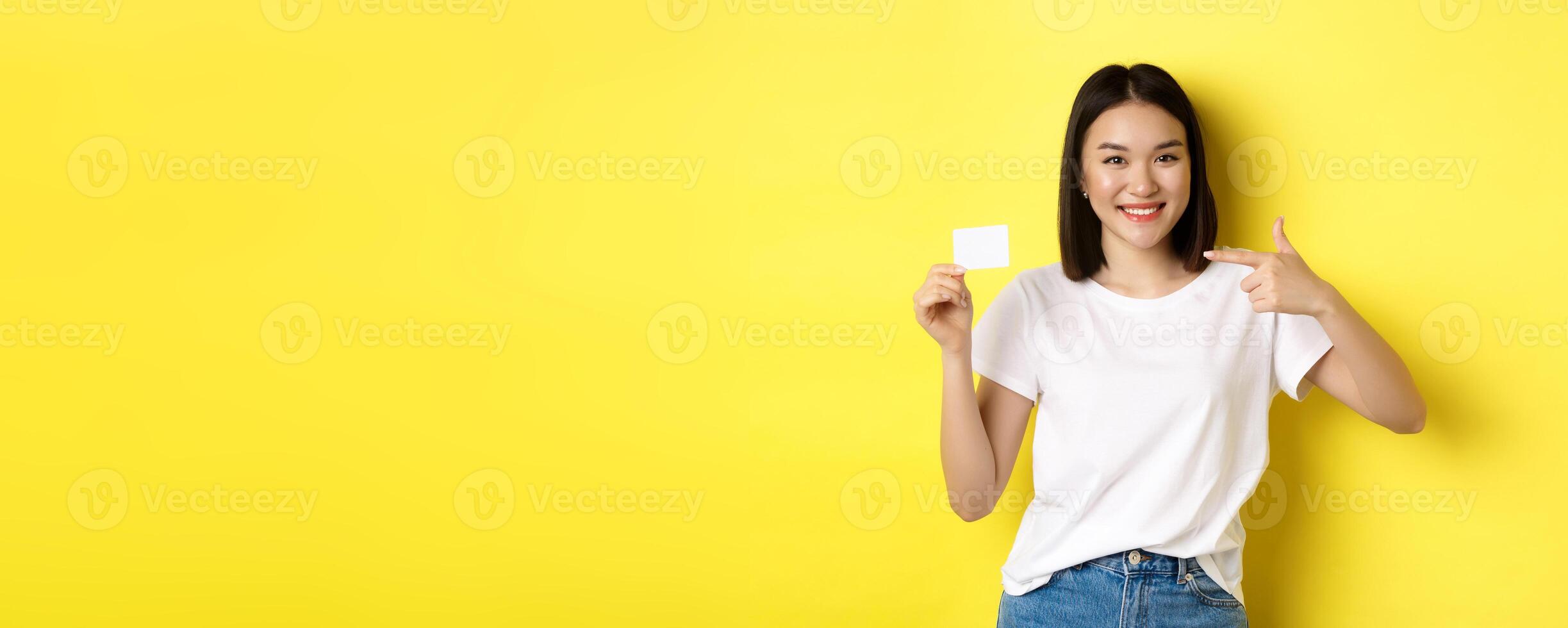 Young asian woman in casual white t-shirt showing plastic credit card and smiling at camera, yellow background photo