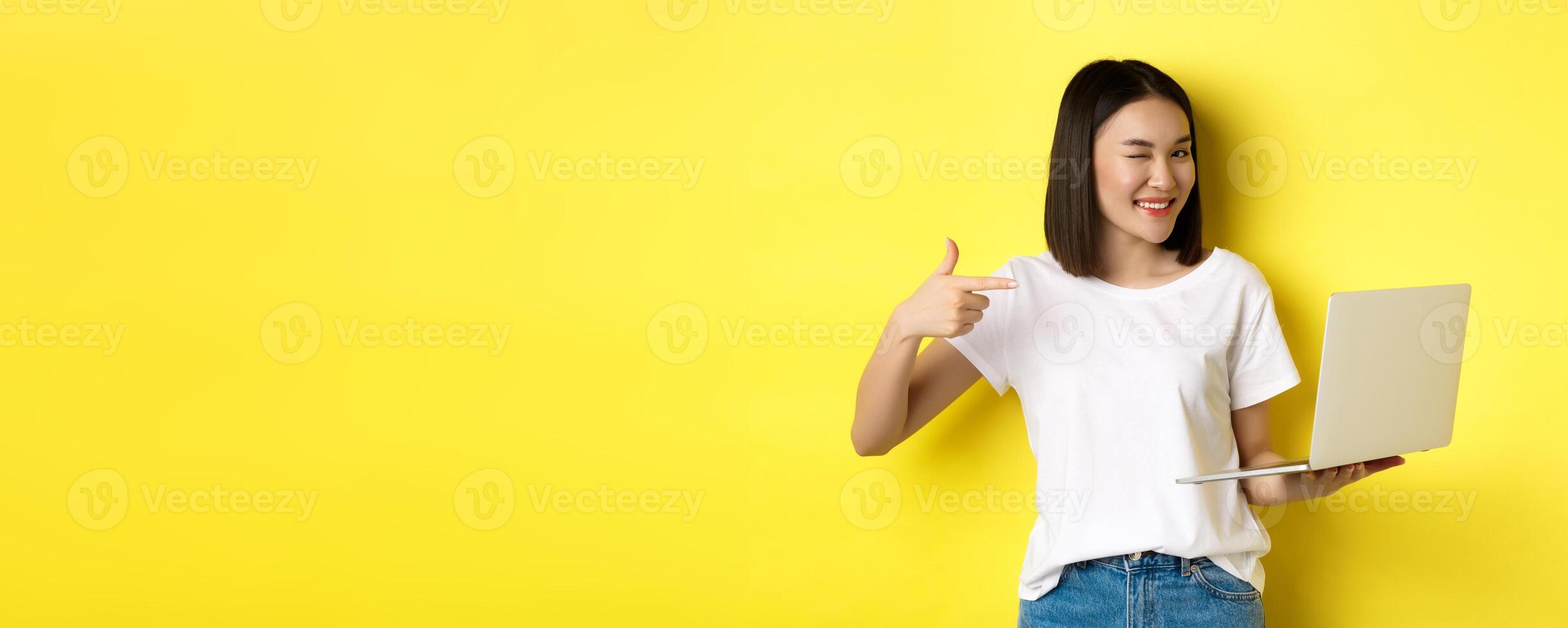 E-commerce and shopping concept. Good-looking asian girl winking at camera, smiling and pointing finger a laptop, showing something, standing over yellow background photo