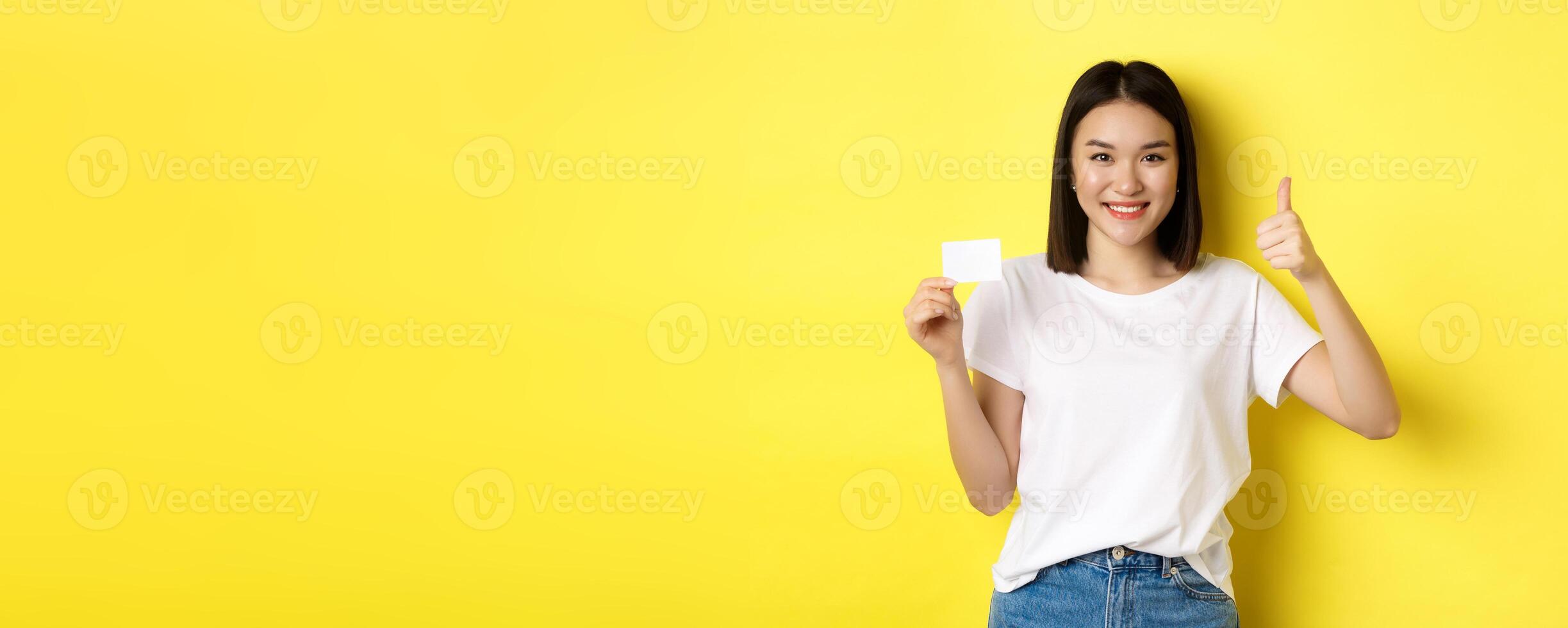 Young asian woman in casual white t-shirt showing plastic credit card and thumb up gesture, approve and recommend, smiling at camera, yellow background photo
