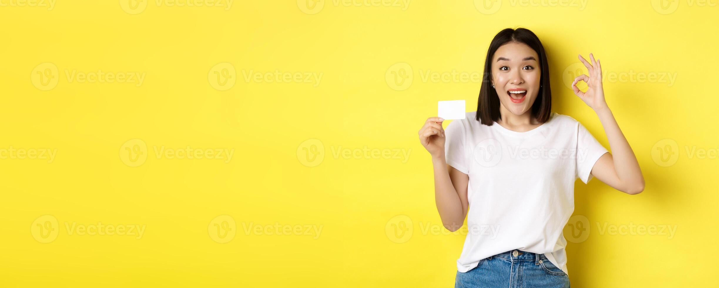Young asian woman in casual white t-shirt showing plastic credit card and okay gesture, recommend bank, smiling at camera, yellow background photo