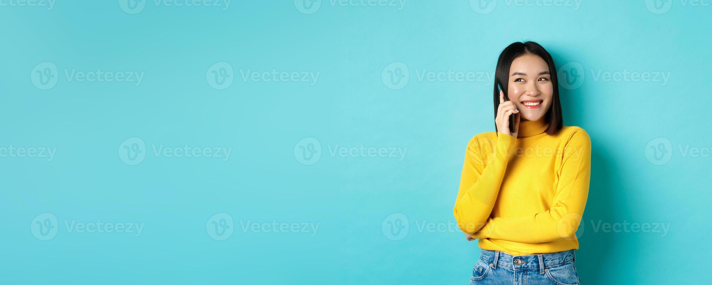 Attractive korean woman in yellow sweater, having conversation and smiling, talking on mobile phone, looking left at copy space, standing over blue background photo