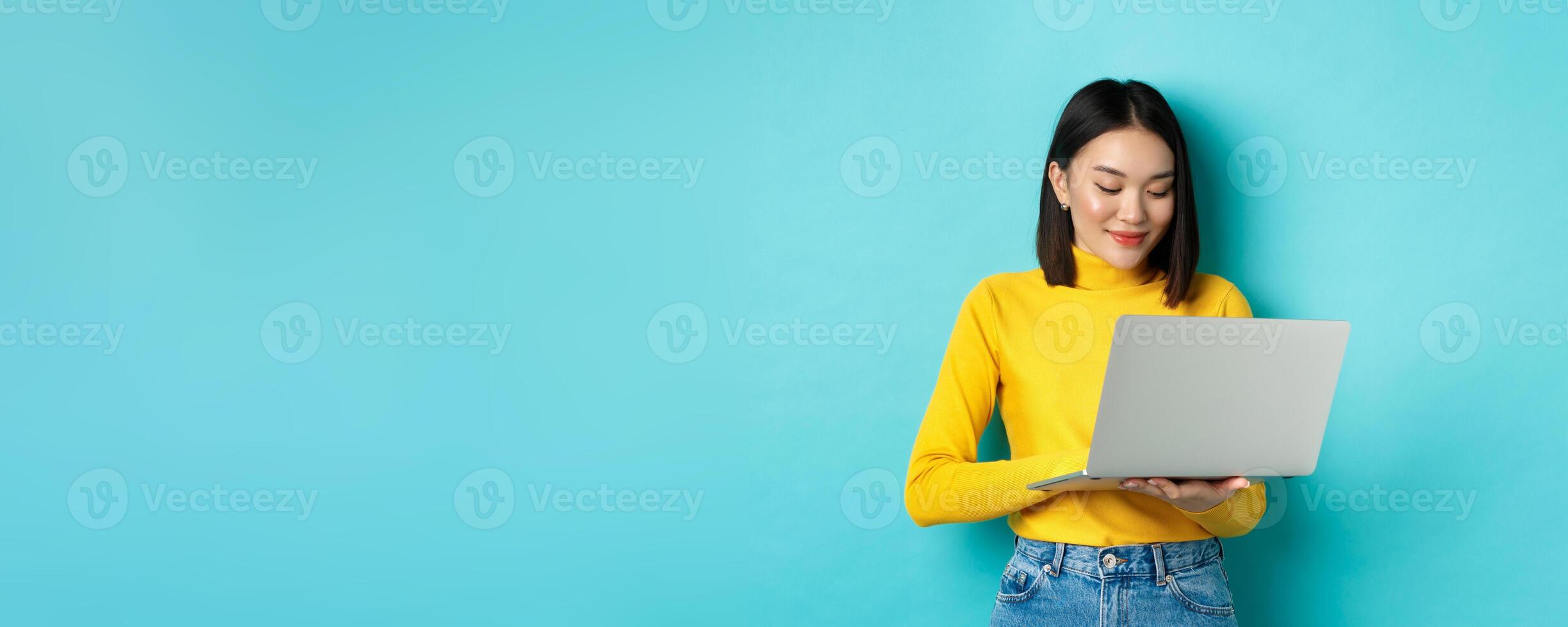 Beautiful asian female student working on laptop, typing on keyboard and looking at screen with pleased smile, standing over blue background photo