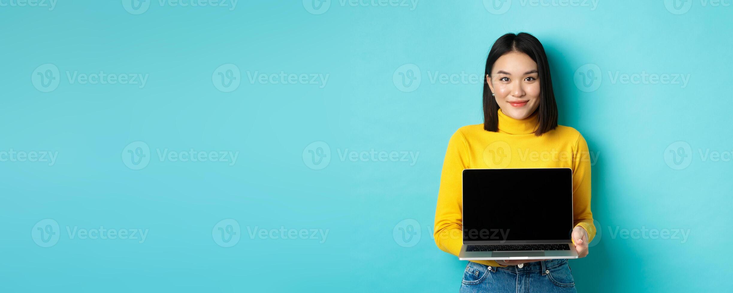 Beautiful and stylish asian woman demonstrate product on screen, showing empty laptop display and smiling, standing over blue background photo