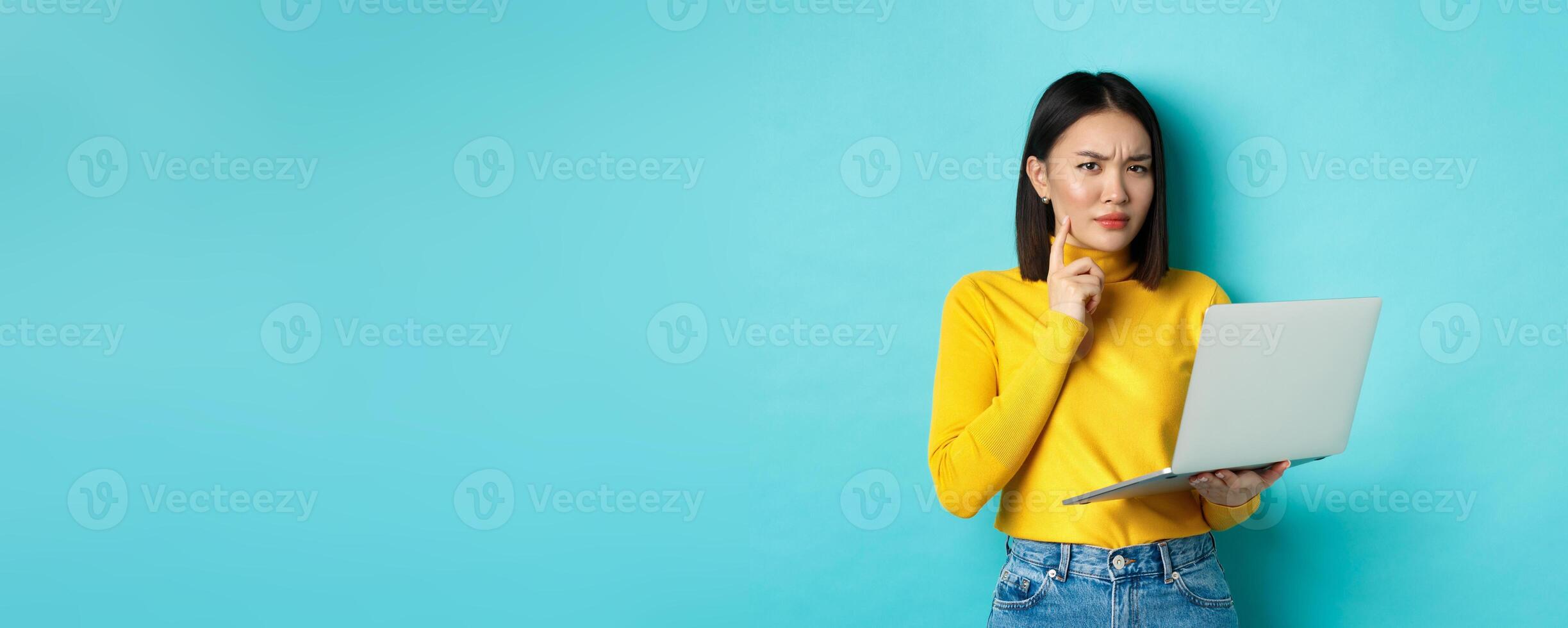 Serious looking asian woman working on laptop and thinking, frowning at camera, solving problem at work, standing over blue background photo