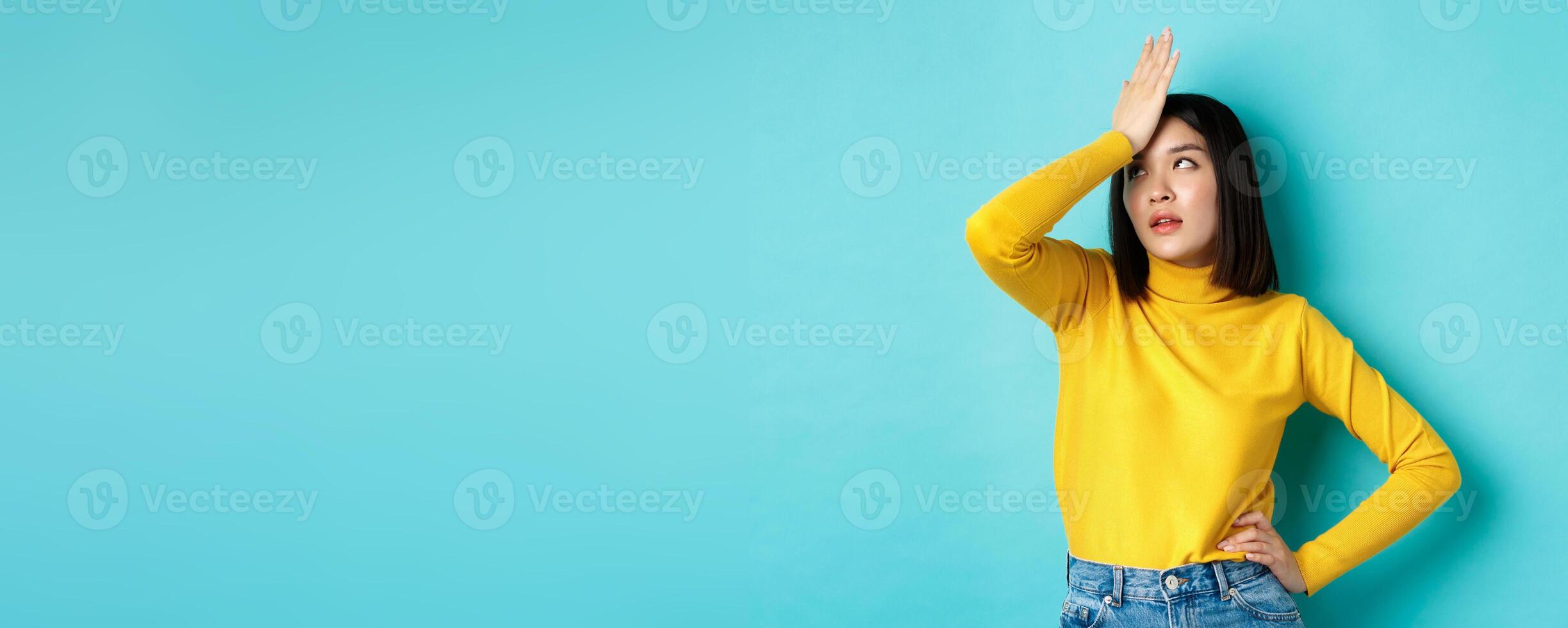 Emotions and lifestyle concept. Annoyed asian girl roll eyes and face palm, standing bothered in yellow pullover against blue background photo