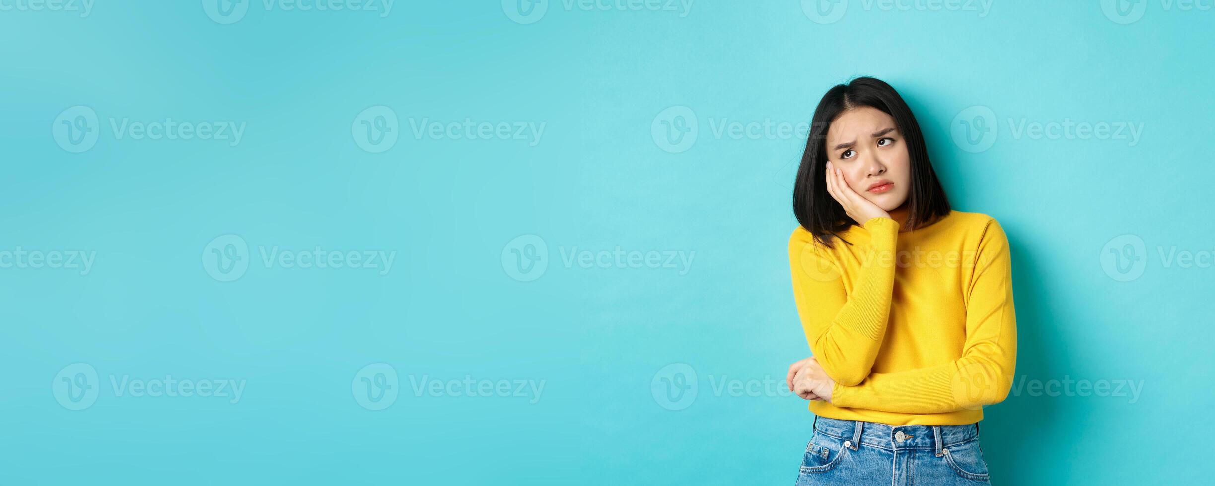 Sad and bored asian teenage girl leaning on palm, looking at upper left corner with lonely and upset expression, standing over blue background photo