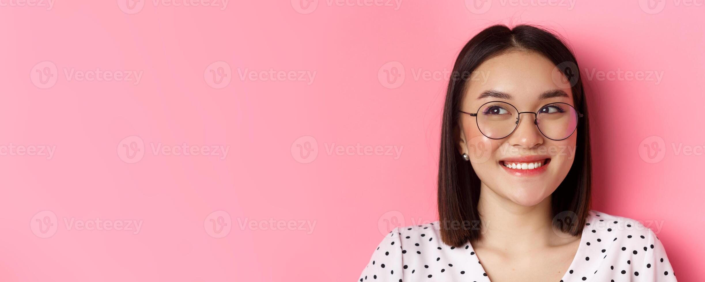 Beauty and lifestyle concept. Close-up of cute asian female model wearing trendy glasses, smiling and looking left at copy space, standing on pink background photo