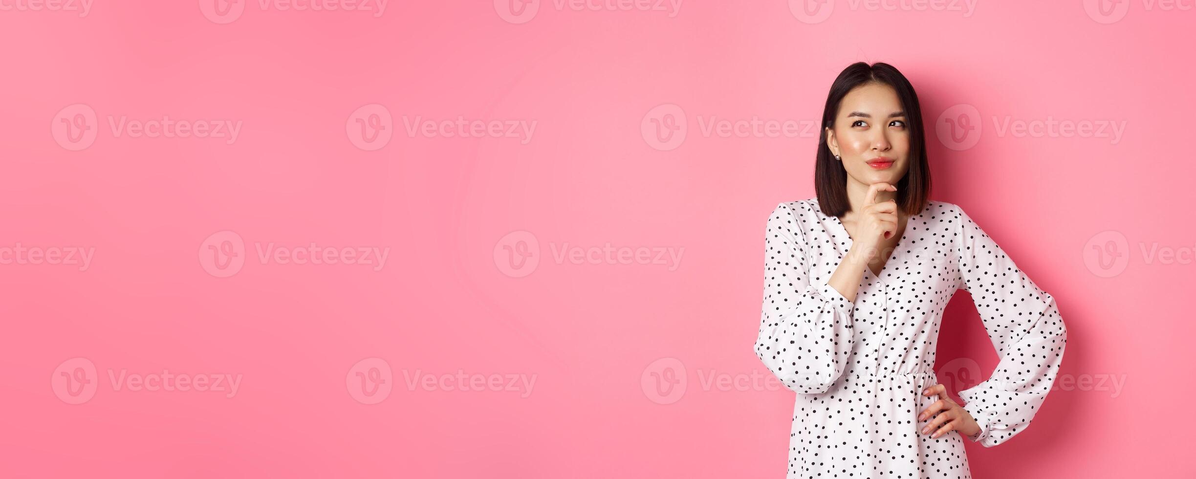 Thoughtful happy asian woman making decision, smiling satisfied and looking at upper left logo, thinking or choosing, standing in dress against pink background photo