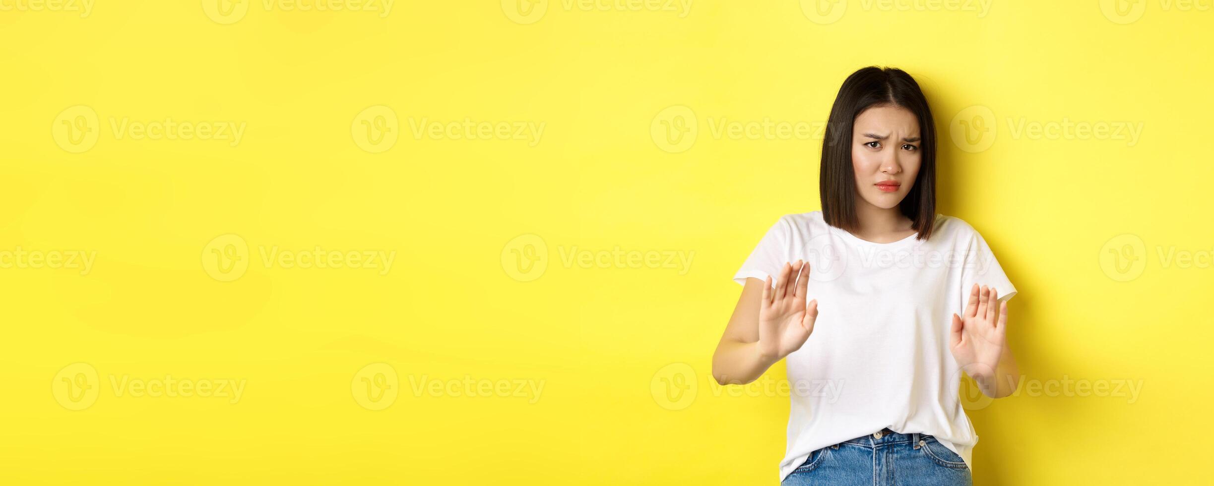 Please no. Asian woman, victim of assualt or domestic abuse, pleading with hands raised up in defense, frowning sad, begging stop, standing over yellow background photo