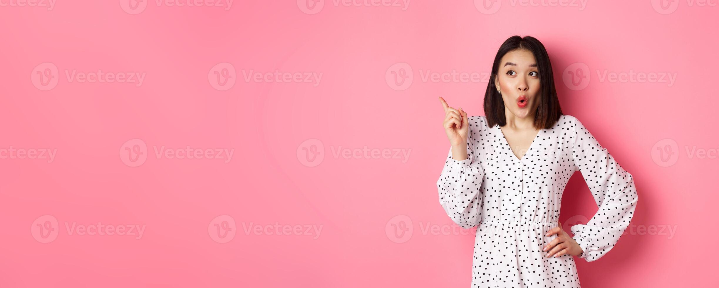 Amazed asian girl in dress pointing finger at upper left corner promo offer, looking interested, shopping and standing over pink background photo