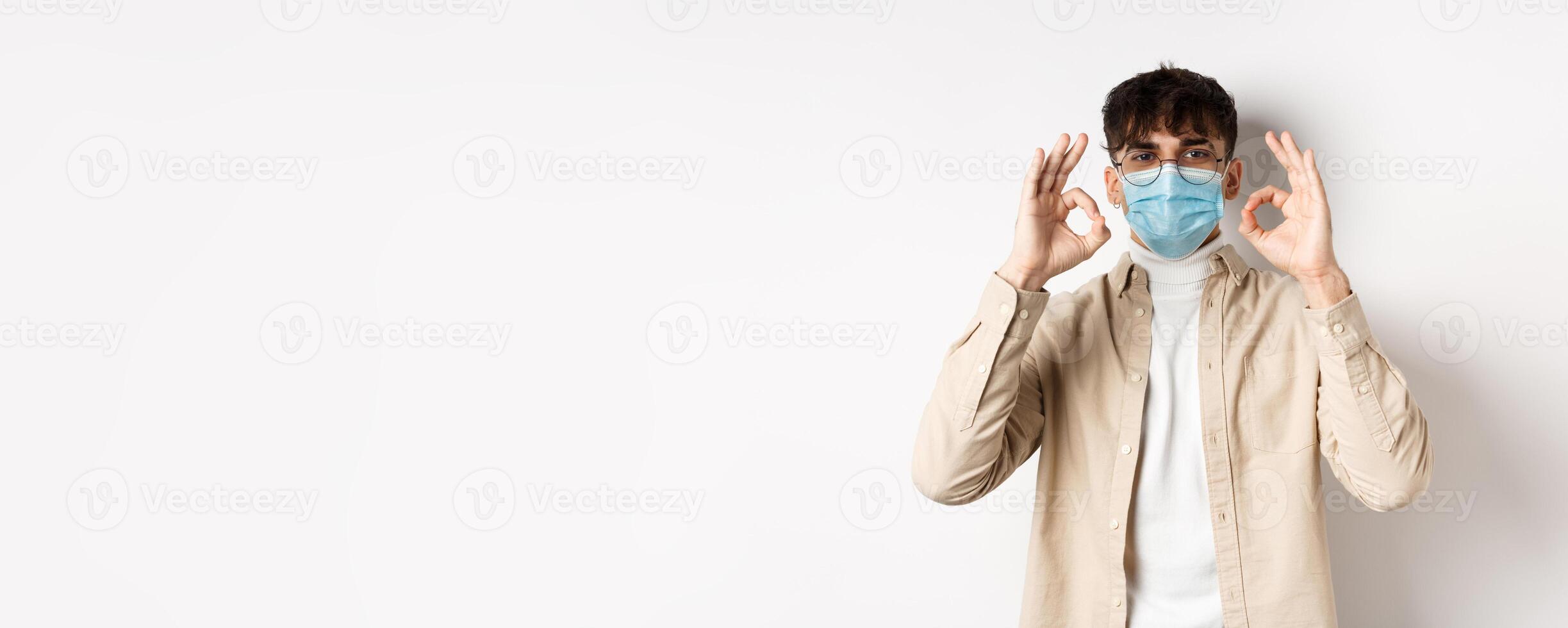 Covid-19, health and real people concept. Handsome guy in glasses and medical mask showing okay gesture in approval, recommend use preventive measures from corona, white background photo