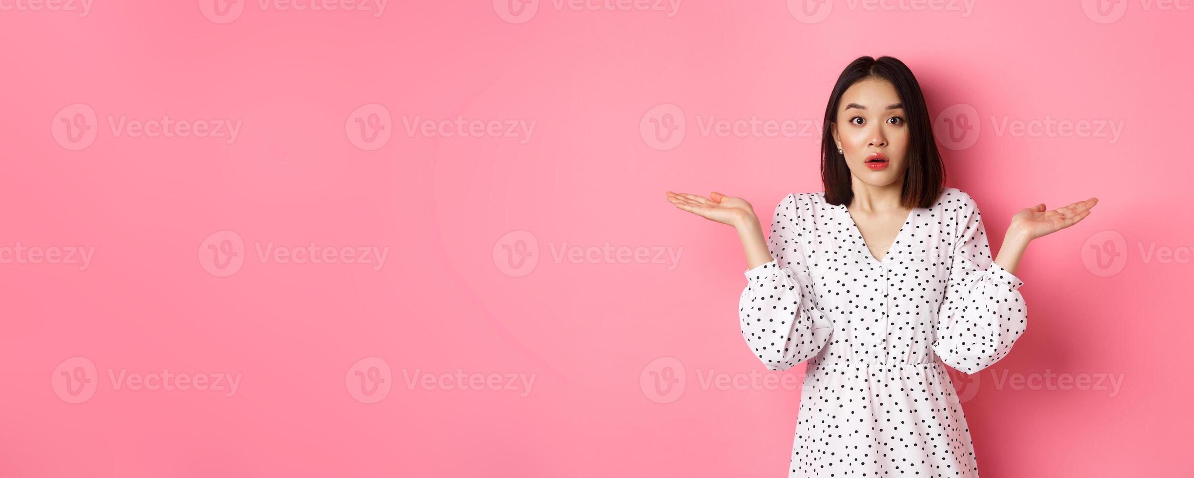 Confused and clueless asian girl shrugging, staring at camera puzzled, dont know what to do, spread hands sideways, standing over pink background photo
