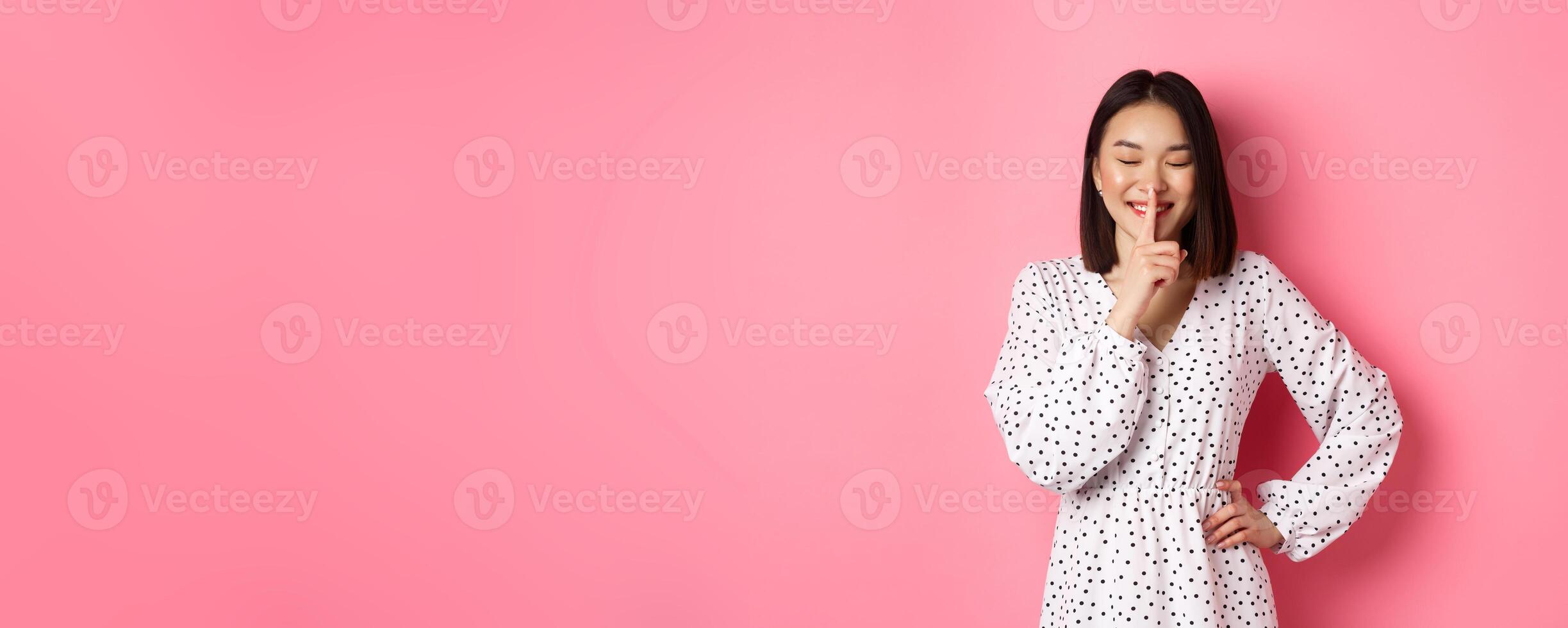 Beautiful Korean woman in trendy dress asking to keep secret, hushing with soft smile and closed eyes, standing over pink background photo