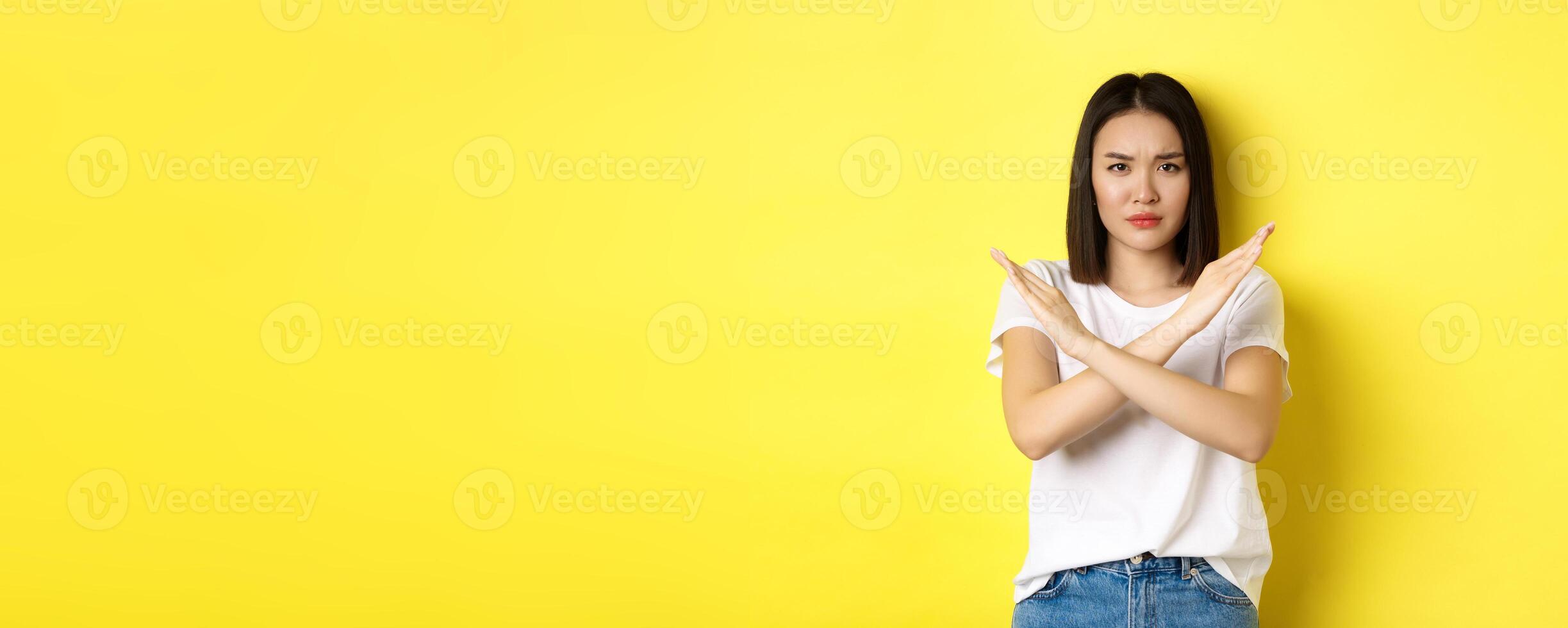 Serious and assertive korean woman showing cross stop gesture, frowning and telling no, prohibit action, disapprove something bad, standing over yellow background photo