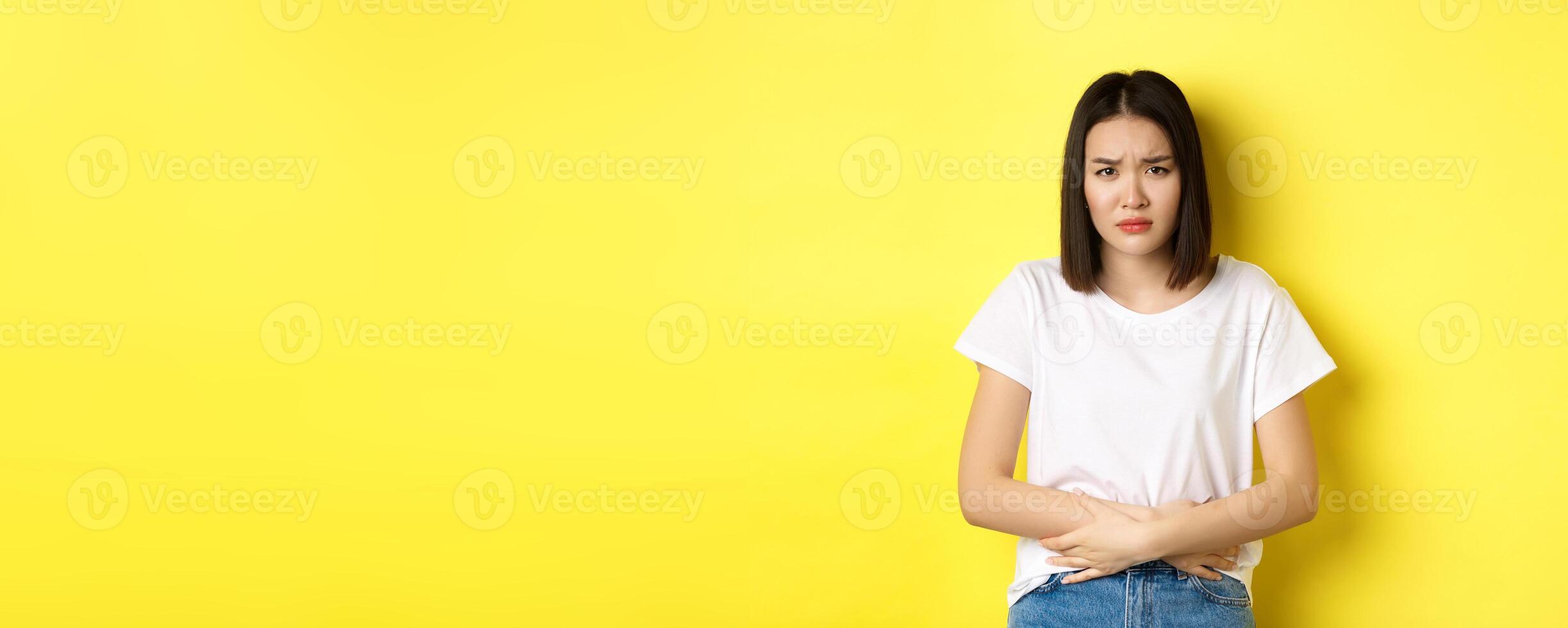 Image of asian woman feeling sick, having painful cramps, holding hands on belly and frowning from pain, discomfort during menstrual period, standing over yellow background photo