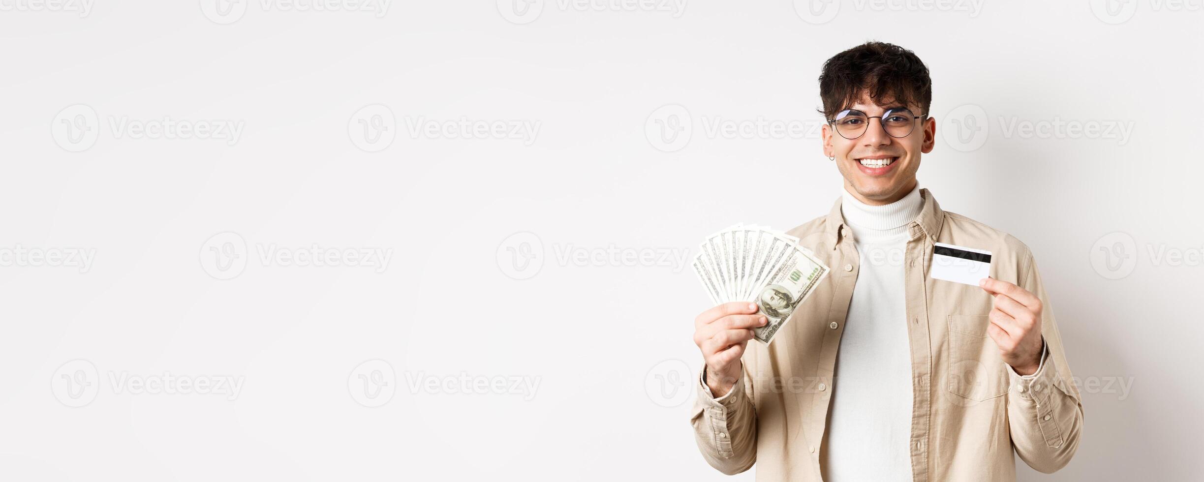 Happy person stands with money and plastic credit card. Young man showing cash and smiling satisfied, standing on white background photo