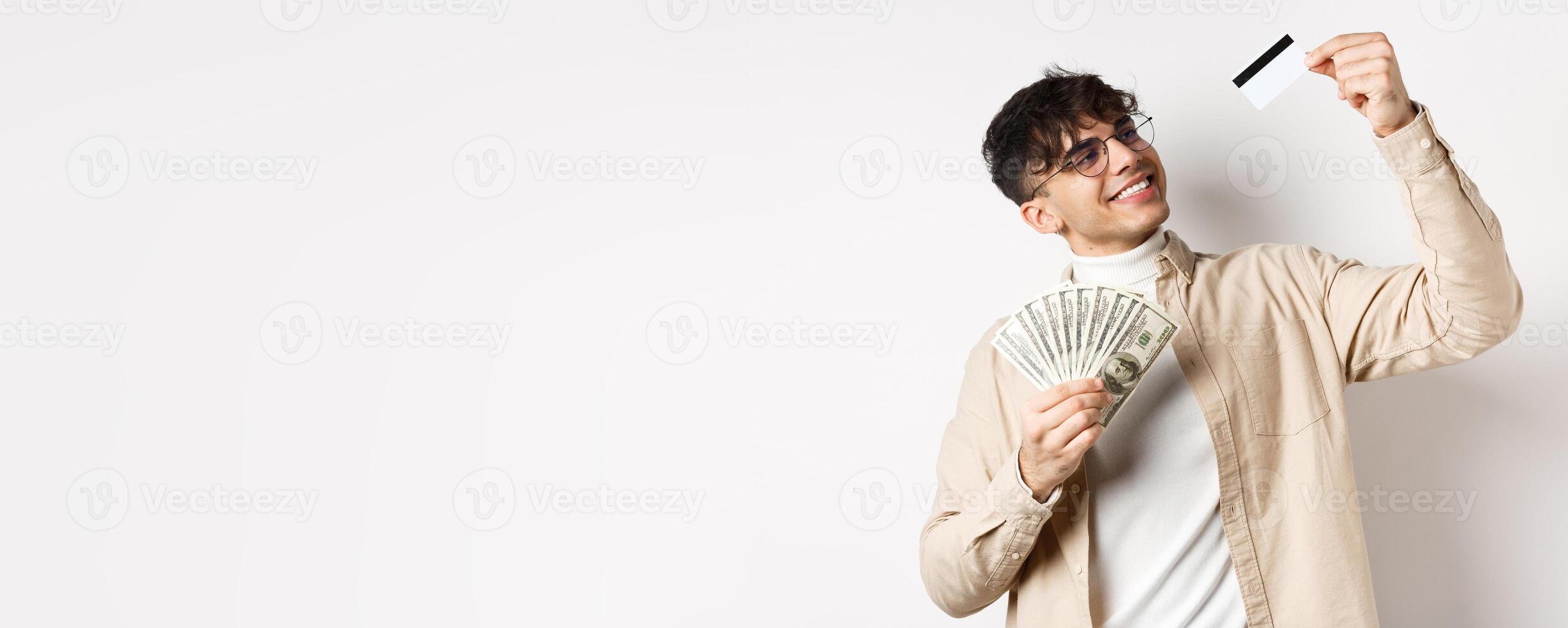 Satisfied young guy holding money, looking pleased and smiling at plastic credit card, choose contactless payment, standing on white background photo
