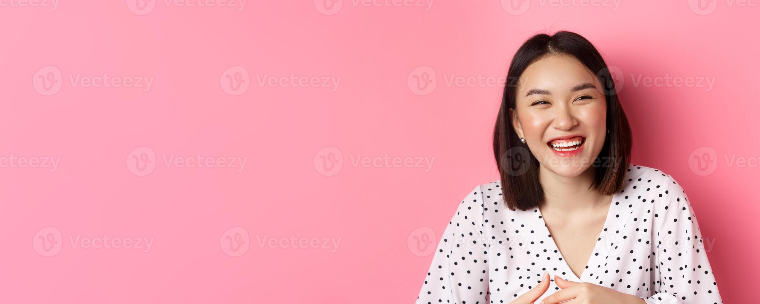 Beauty and lifestyle concept. Close-up of happy asian woman laughing and having fun, standing over pink background photo