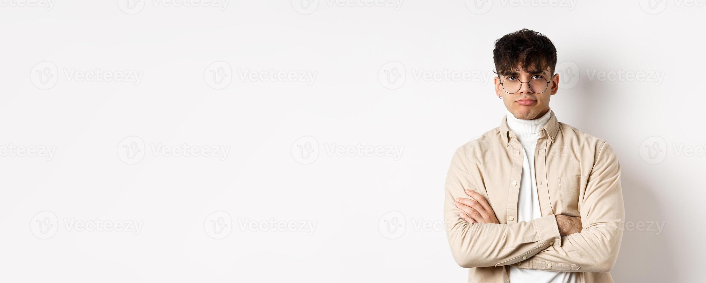 Image of stylish hipster guy in glasses feeling bored, looking unamused with pokerface, cross arms on chest and stand reluctant against white background photo