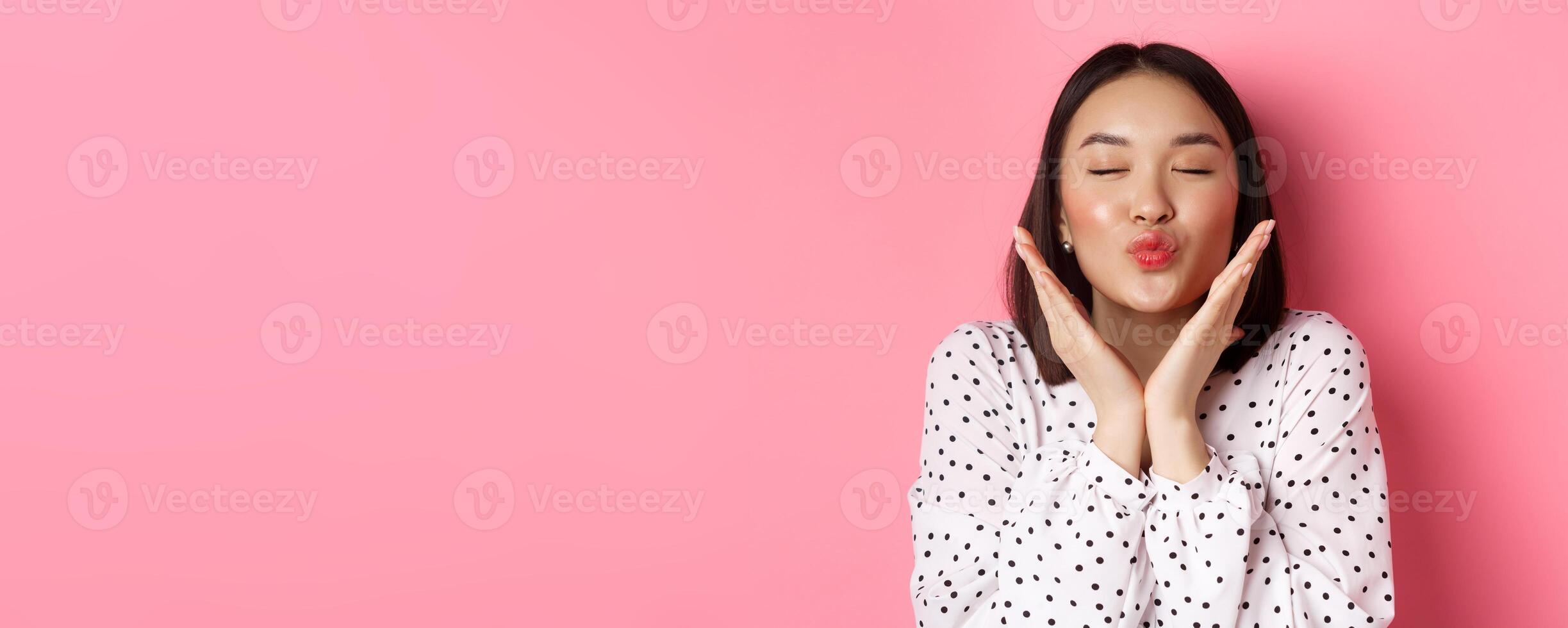 Beauty and skin care concept. Close-up of beautiful asian girl close eyes, pucker lips for kisses, showing cute perfect face, standing over pink background photo