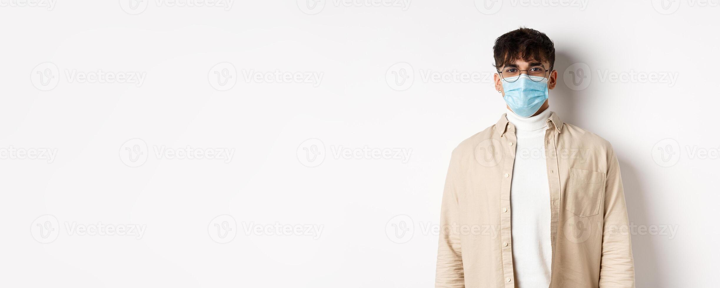 Young man in face mask from covid-19 looking cheerful at camera, standing on white background. Coronavirus, quarantine and social distancing concept photo