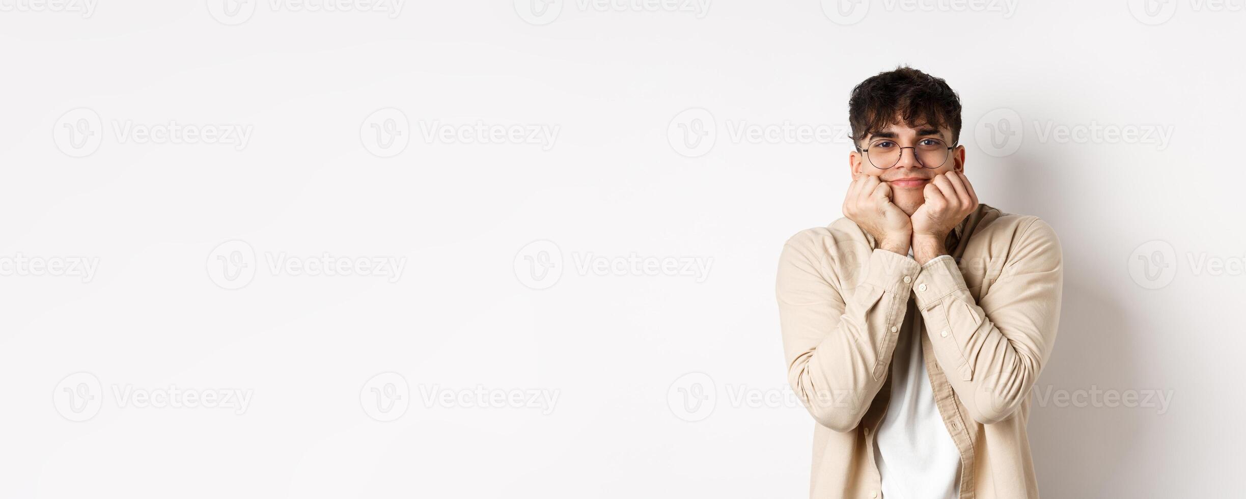 Portrait of handsome caucasian man watching something lovely and cute, squeeze cheeks and lean on hands, looking with admiration and interest at camera, standing on white background photo