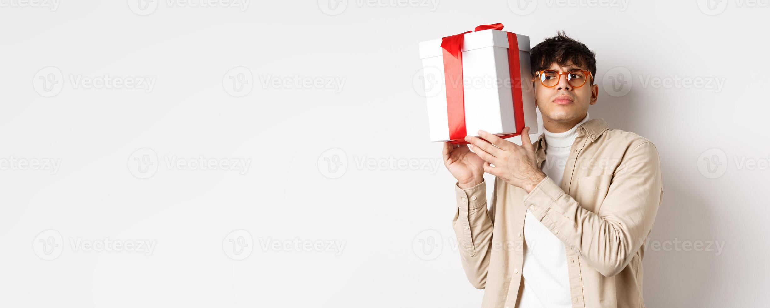 Intrigued guy listening whats inside gift box, trying guess present, standing focused on white background photo
