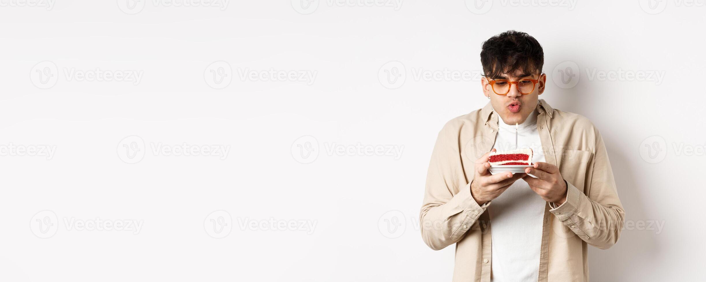 Funny young guy in glasses blowing candle and making wish on birthday cake, standing cheerful against white background photo