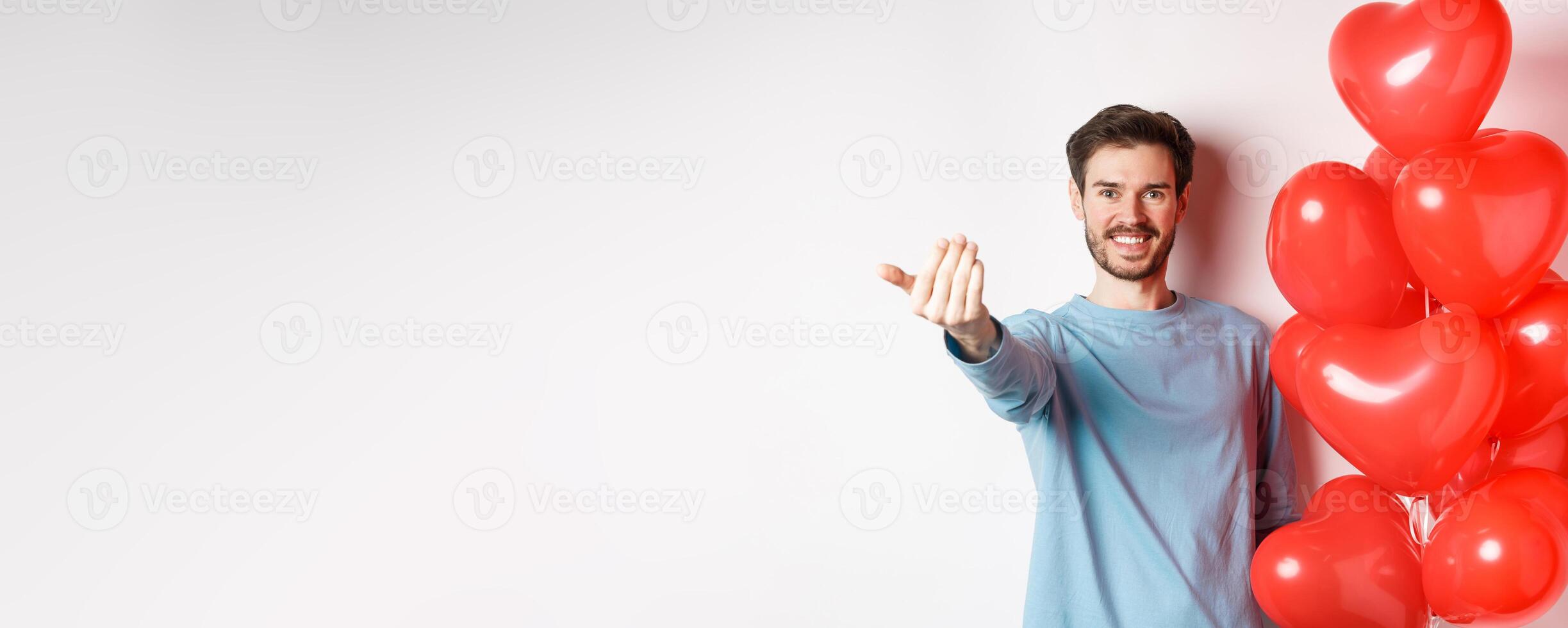 Smiling man beckon you to come closer, follow me gesture, taunting his lover move forward, have romantic surprise, standing near red balloon on valentines day, white background photo