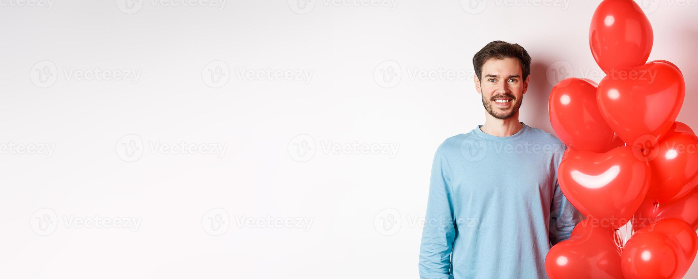 Smiling young man standing with heart balloons and looking happy, celebrating valentines day, bring romantic present to lover, standing over white background photo