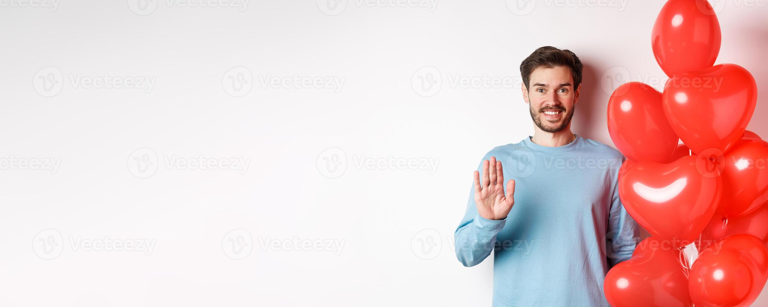 Handsome boyfriend saying hello, bring romantic red heart balloons on date, waving hand and smiling, standing over white background photo