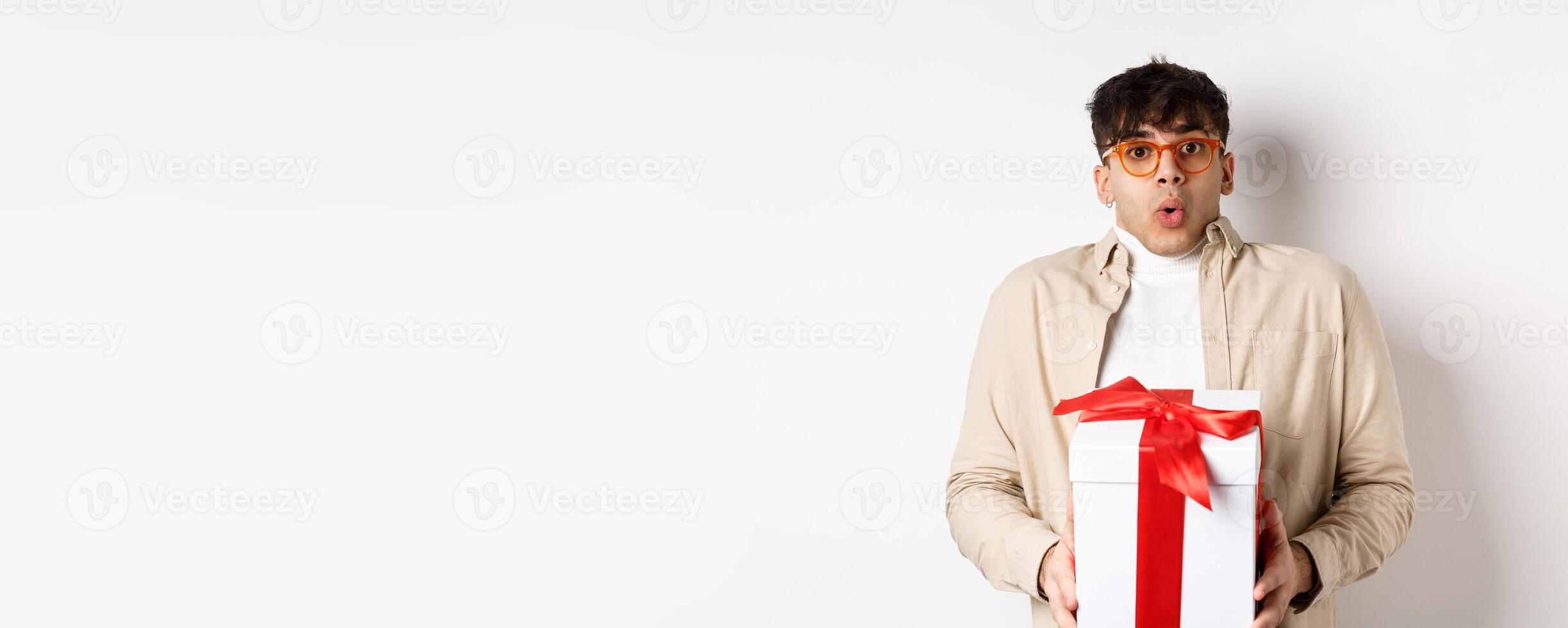 Surprised young man say wow and holding gift box, receive present on holiday, standing amused on white background photo