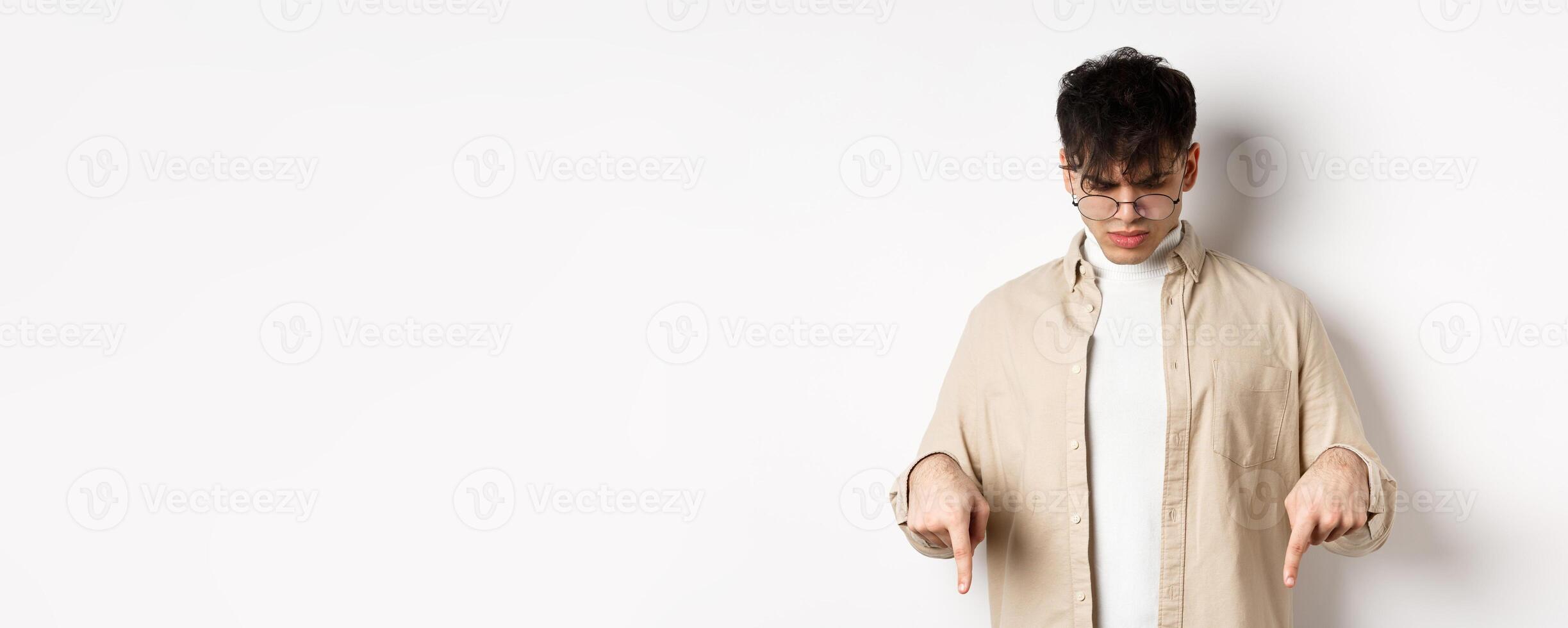 Confused frowning guy in glasses looking, pointing fingers down at something strange, standing puzzled on white background photo