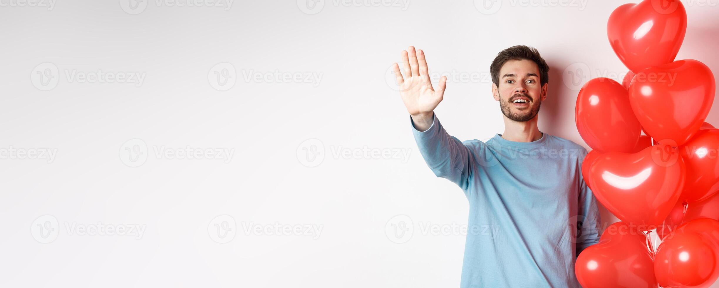 Handsome young caucasian man standing with romantic heart balloon and waving hand at lover, waiting for his date on Valentines day with cute surprise, standing over white background photo