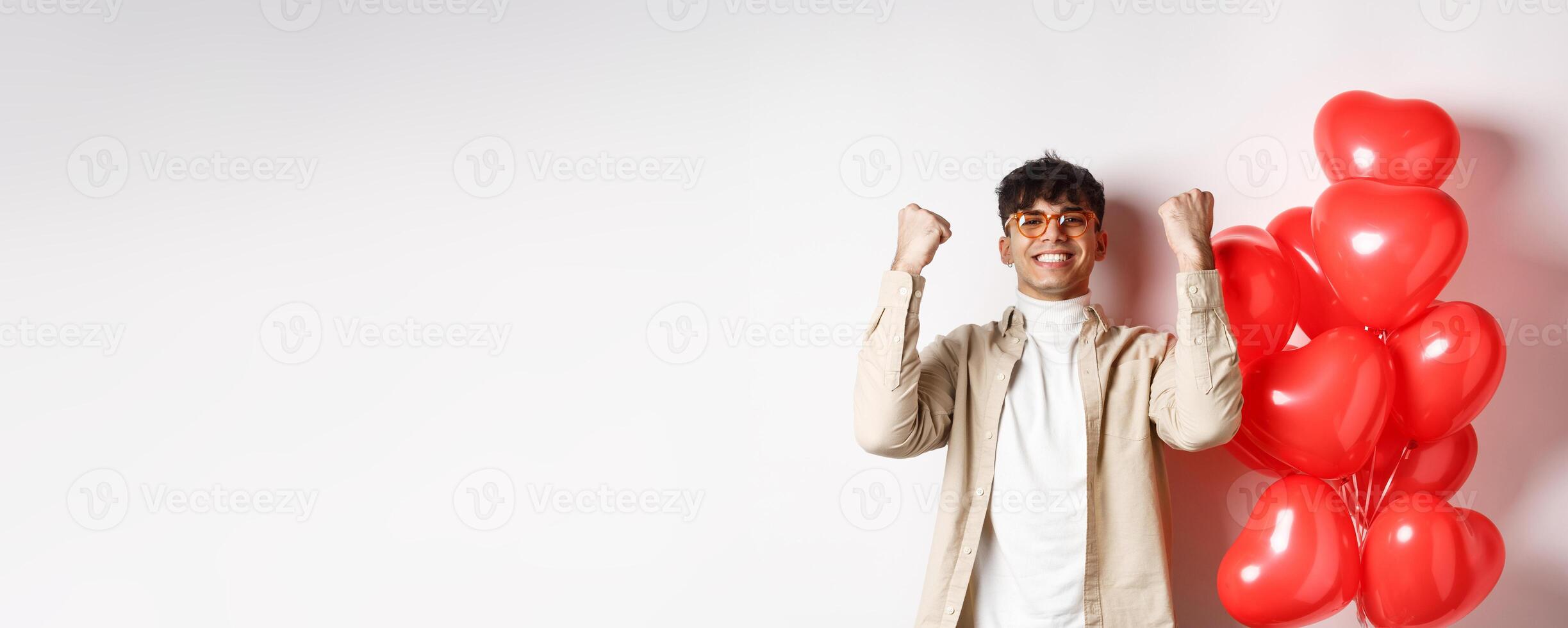 Valentines day. Satisfied young man saying yes, triumphing and celebrating on lovers date, making fist pump and smiling pleased, standing near heart balloons on white background photo