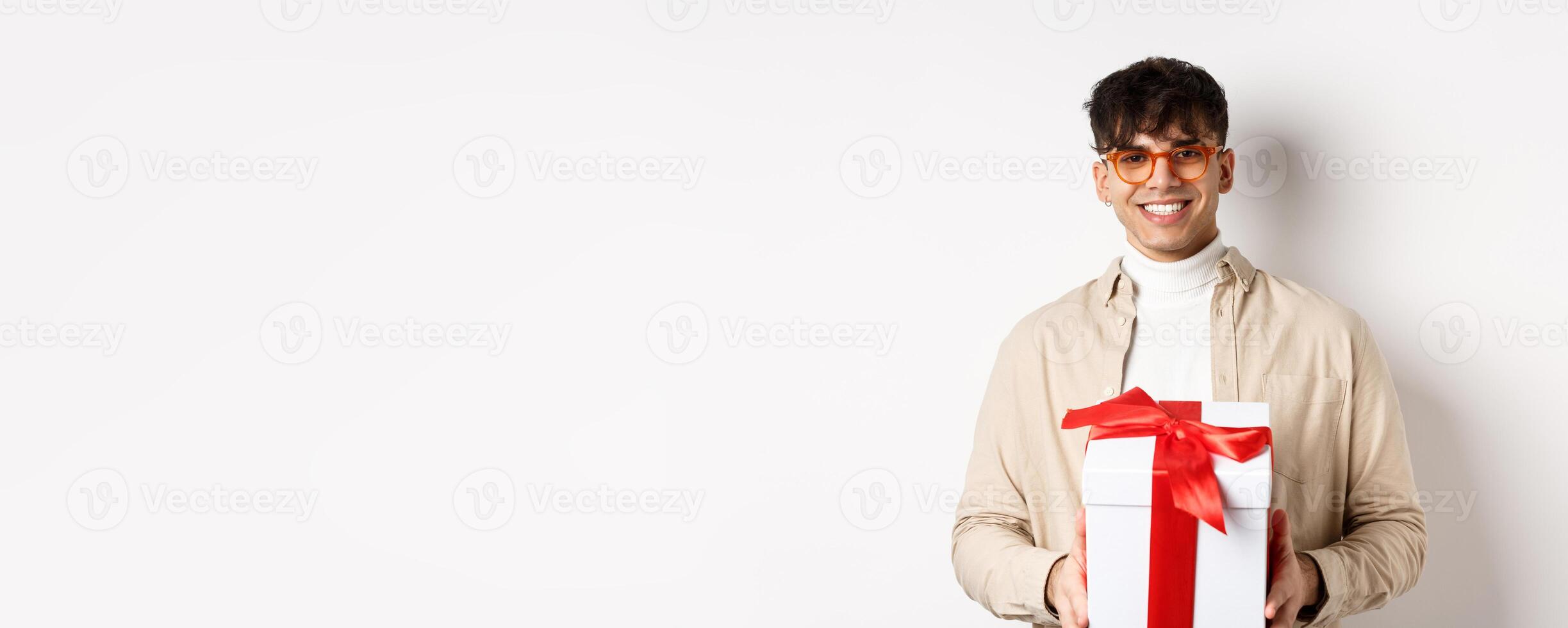 Handsome young man making a gift, holding box with present and smiling at camera, standing on white background photo