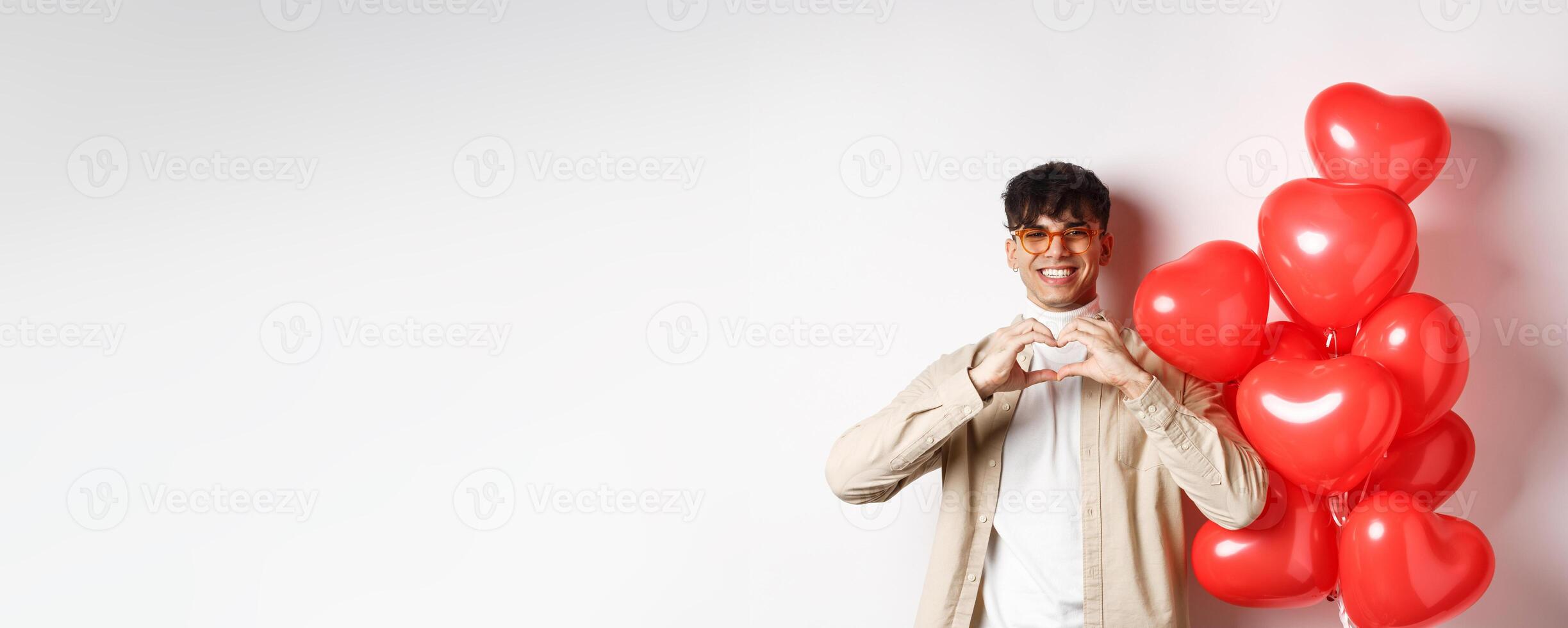 Valentines day and romance concept. Handsome young man standing near red hearts balloons and showing heart gesture, being in love and smiling, white background photo