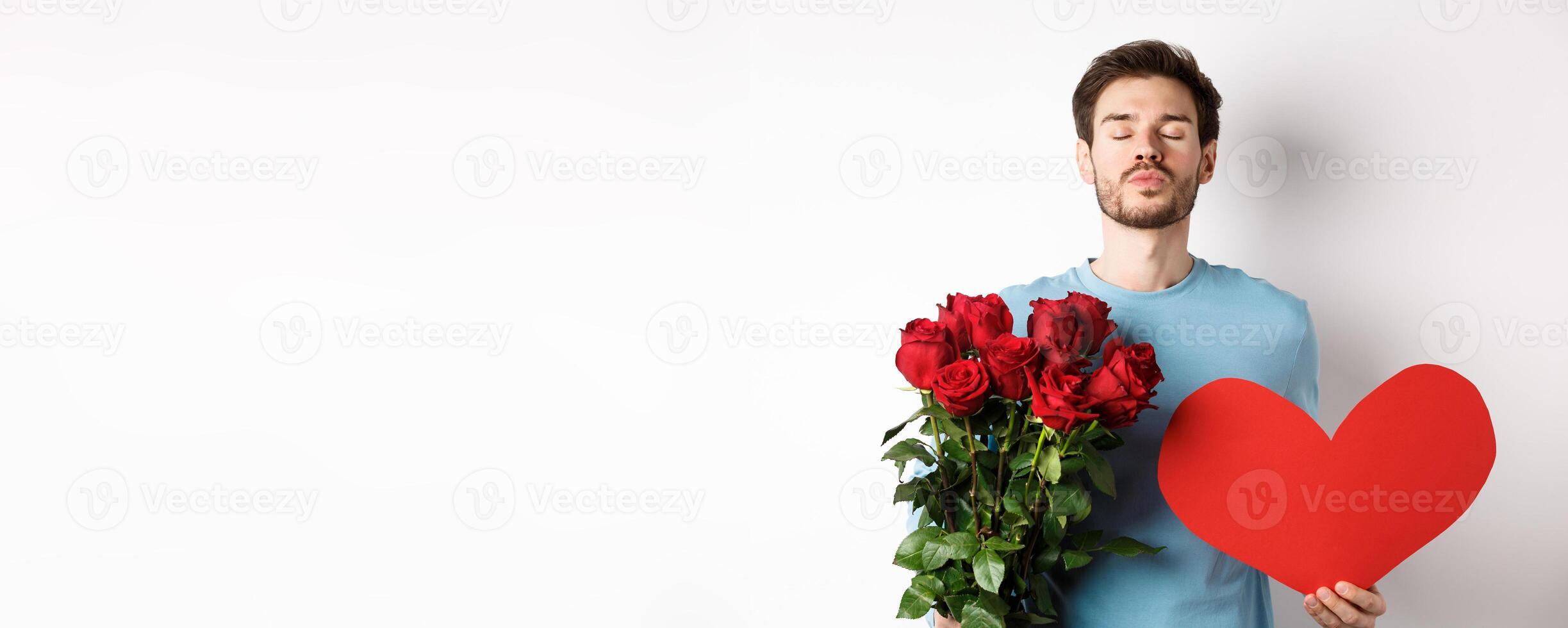 Romantic boyfriend waiting for kiss, holding bouquet of roses flowers and big red heart on Valentines day, love in air, standing over white background photo