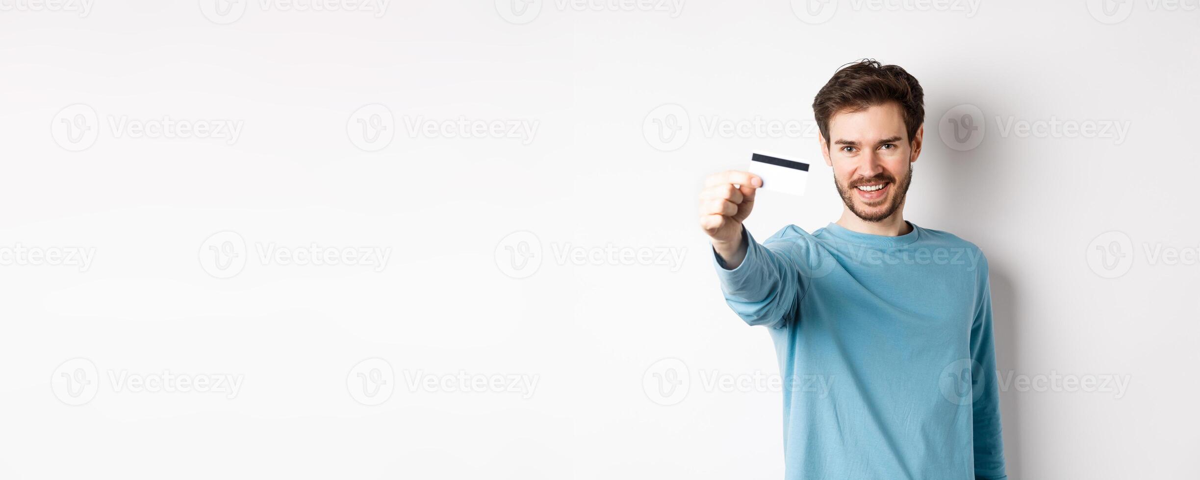confidente joven hombre sonriente, tramo fuera mano y demostración el plastico crédito tarjeta, en pie en blanco antecedentes foto