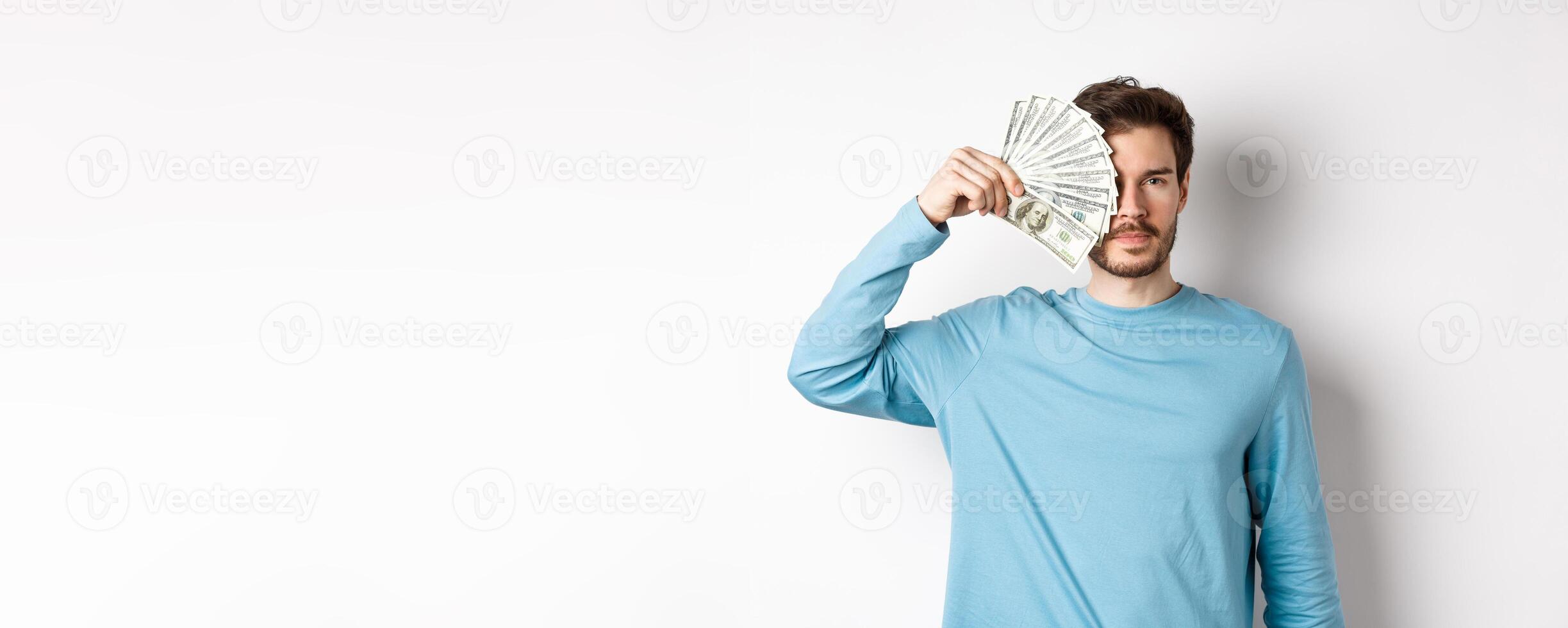 Serious young man covering half of face with money, looking at camera, standing in blue sweatshirt over white background photo