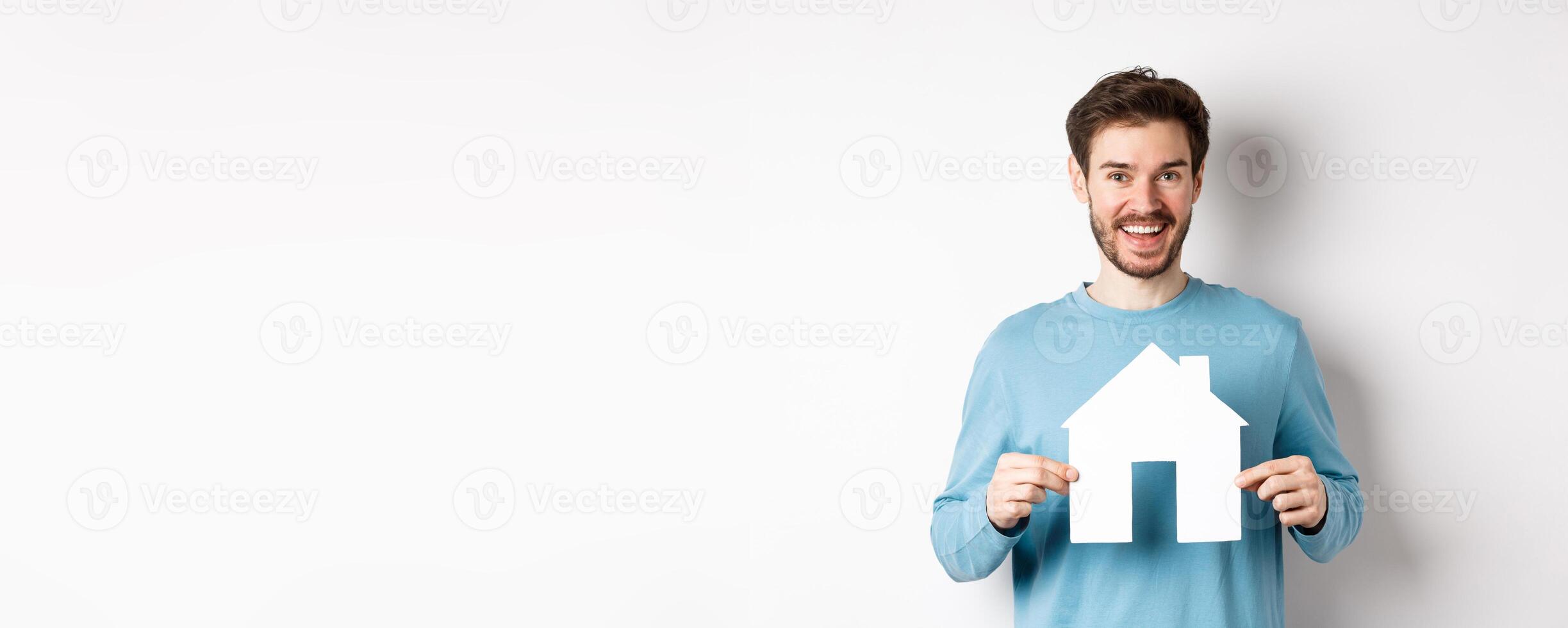 Real estate and insurance concept. Handsome modern man buying property, smiling and showing paper house cutout, standing over white background photo