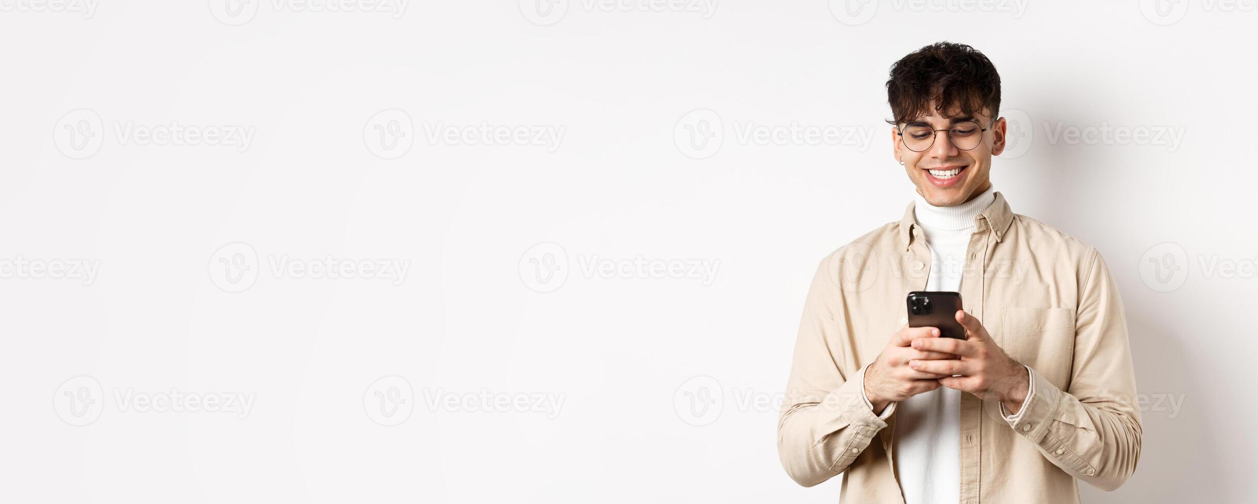Real people. Natural young man reading message on mobile phone, smiling and looking at smartphone screen, standing on white background photo
