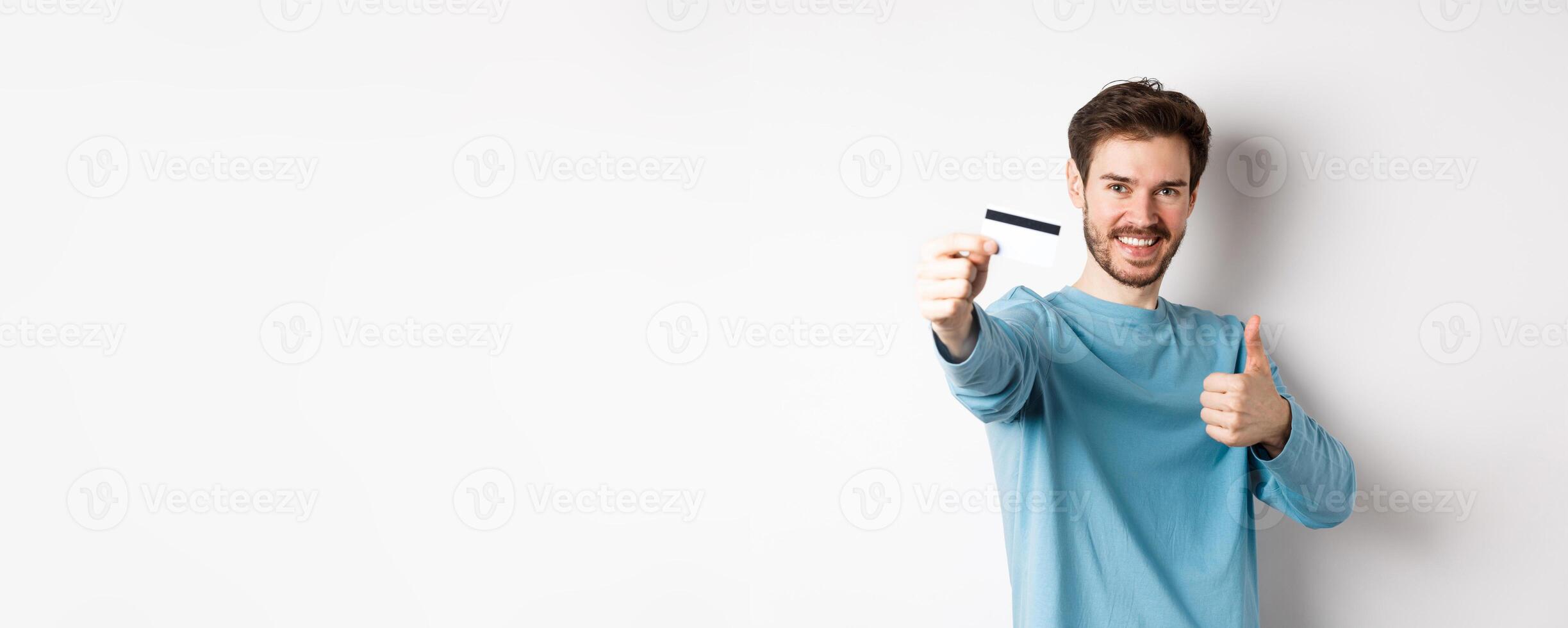 Handsome caucasian man showing plastic credit card with thumb-up, recommend and praise good bank offer, standing on white background. photo