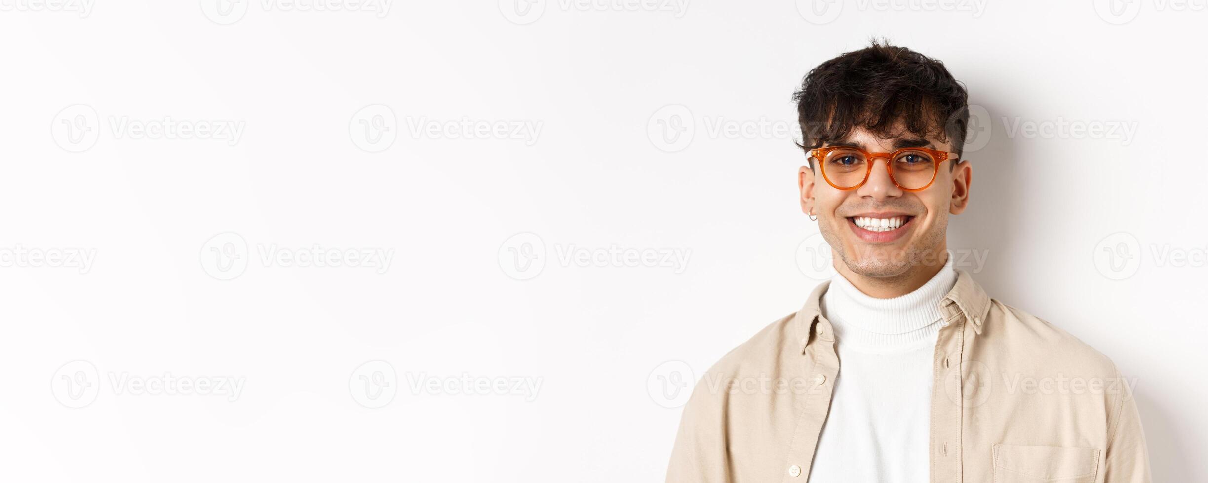 Close-up of handsome hipster guy with natural smile, wearing glasses and earrings, looking happy at camera, standing on white background photo