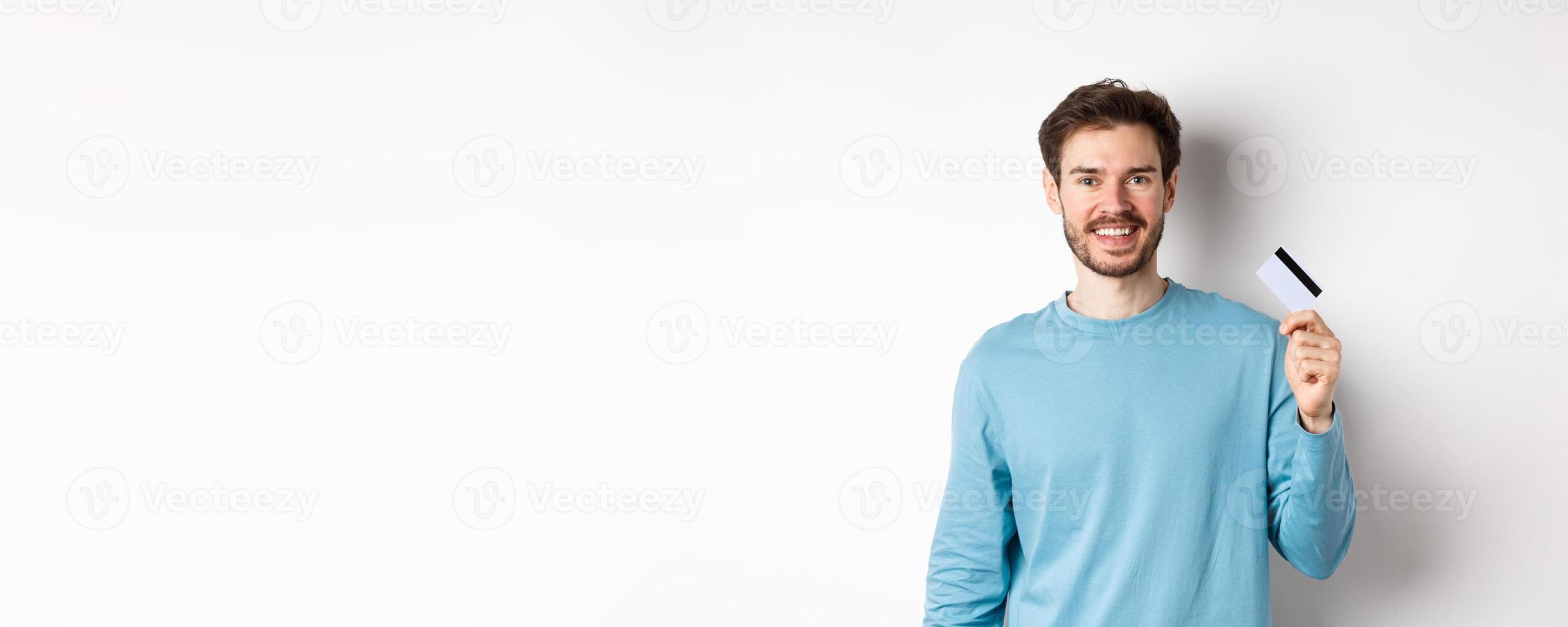 Smiling male bank client showing plastic credit card with happy face, standing over white background photo