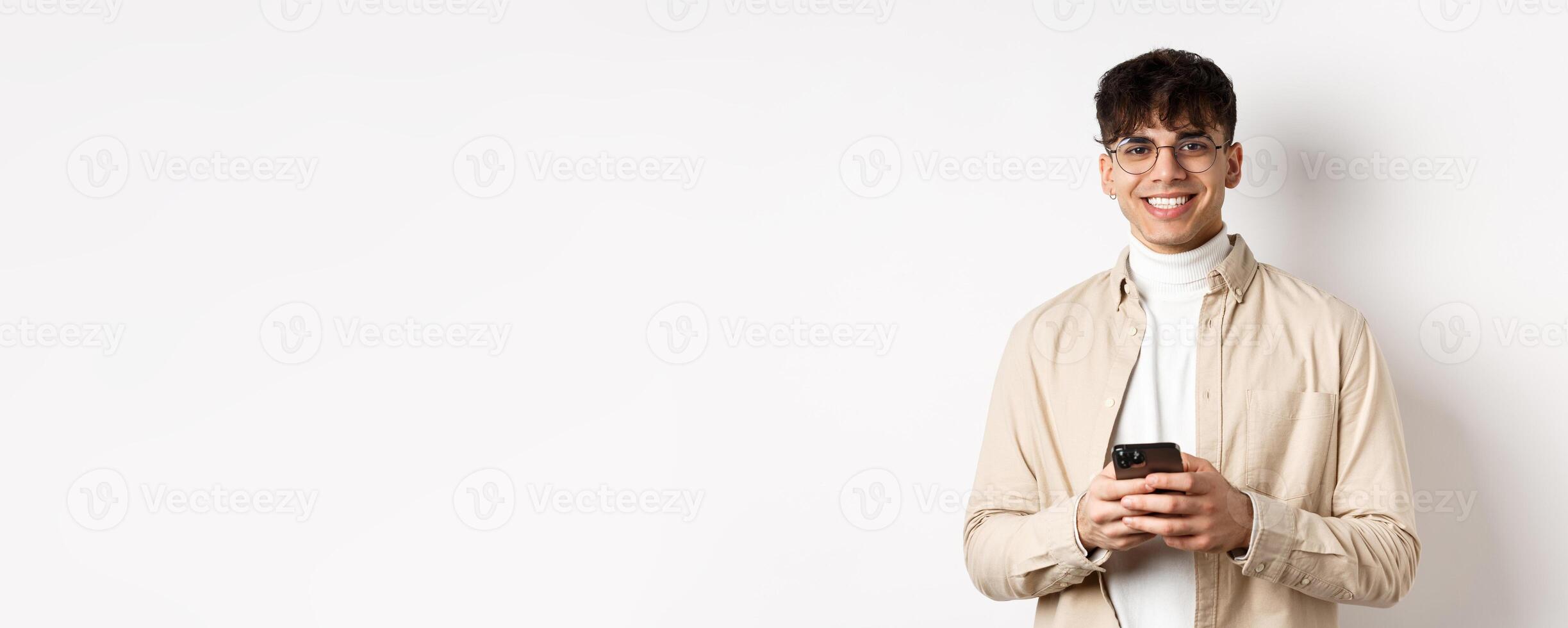Real people. Handsome guy in glasses using mobile phone and smiling happy at camera, standing on white background photo