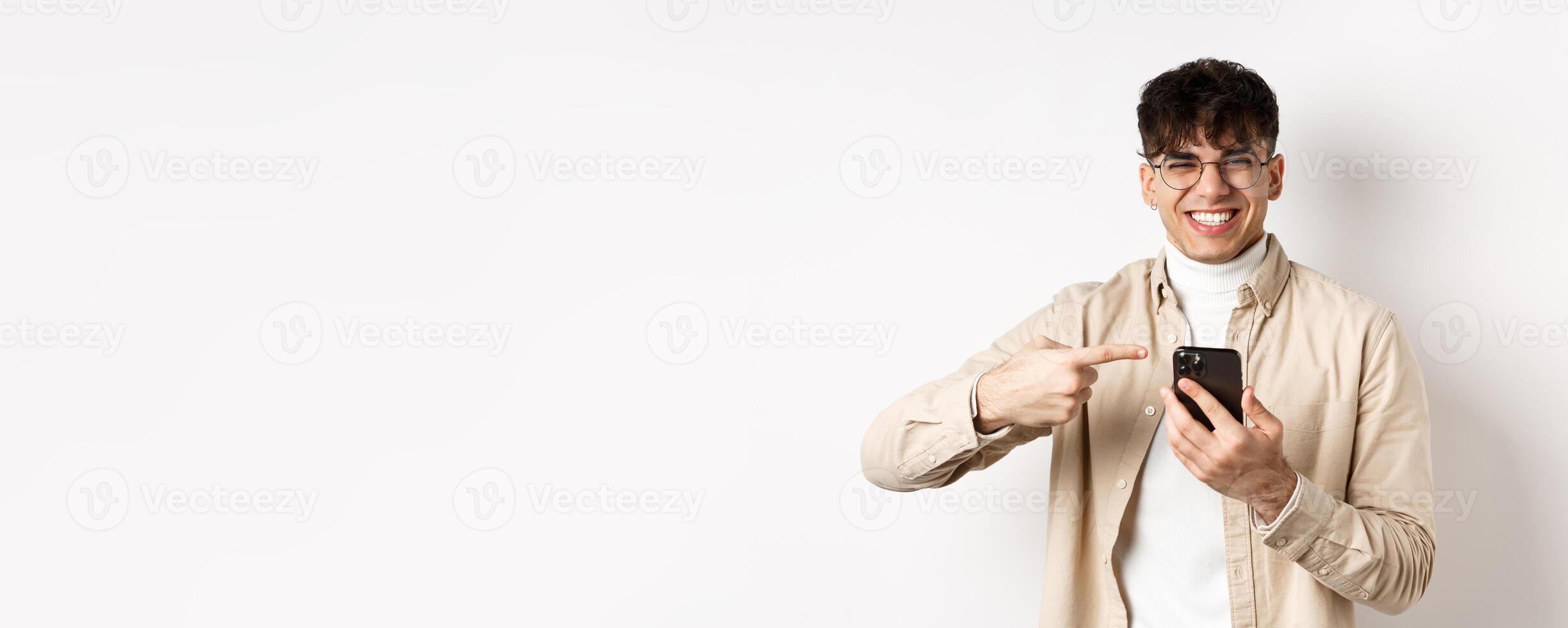 Technology and online shopping concept. Portrait of natural man in glasses laughing and pointing finger at smartphone, showing promo offer on screen, standing white background photo
